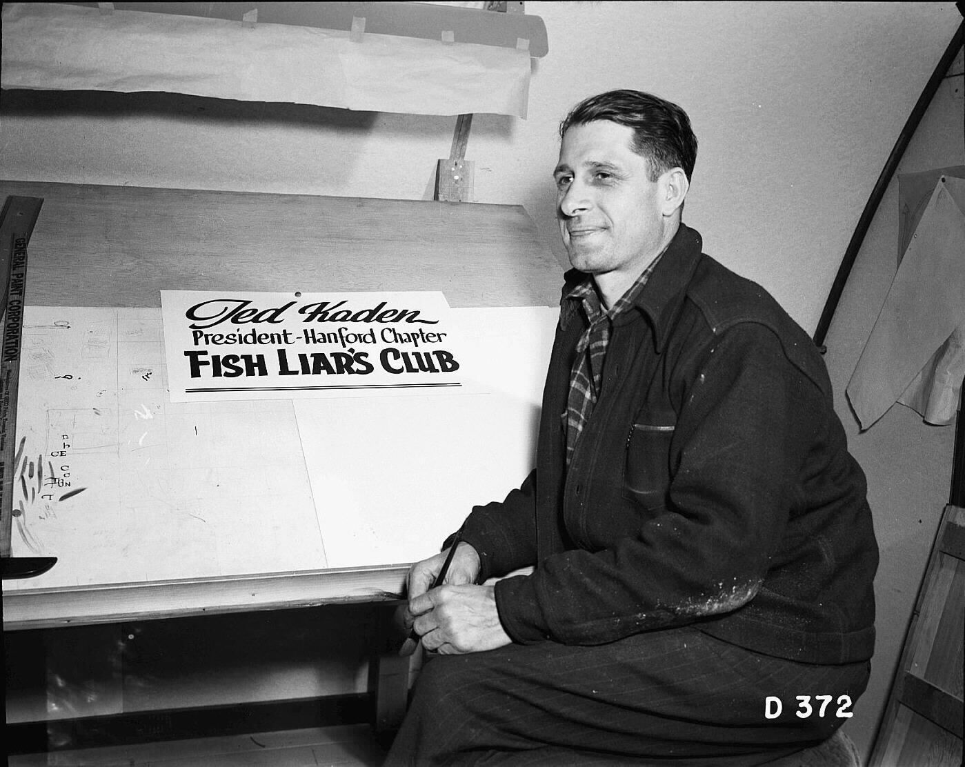 Black and white photo of a man by a desk and a sign that reads "Jed Kaden", "President Hanford Chapter", and "FISH LIAR'S CLUB" in different fonts. 