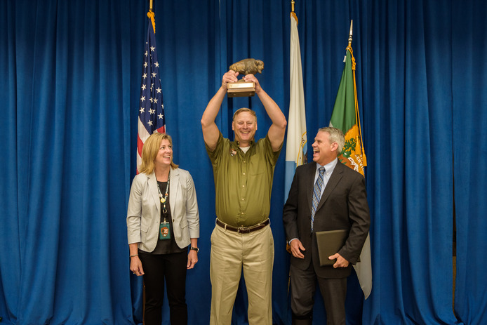 a tall man holds a small bison statue high in the air as he stands in between a man and a woman in business clothes. The background is a blue curtain with an American flag and a National Park Service flag