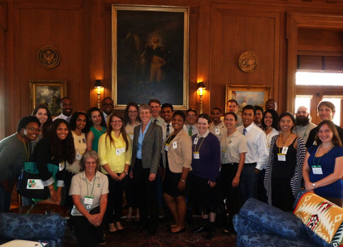 Inside a wood paneled room with large painted portraits hanging on the wall, a group of about 20 people in business casual clothes and ID badges stand on either side of a woman with short gray hair in a suit. 