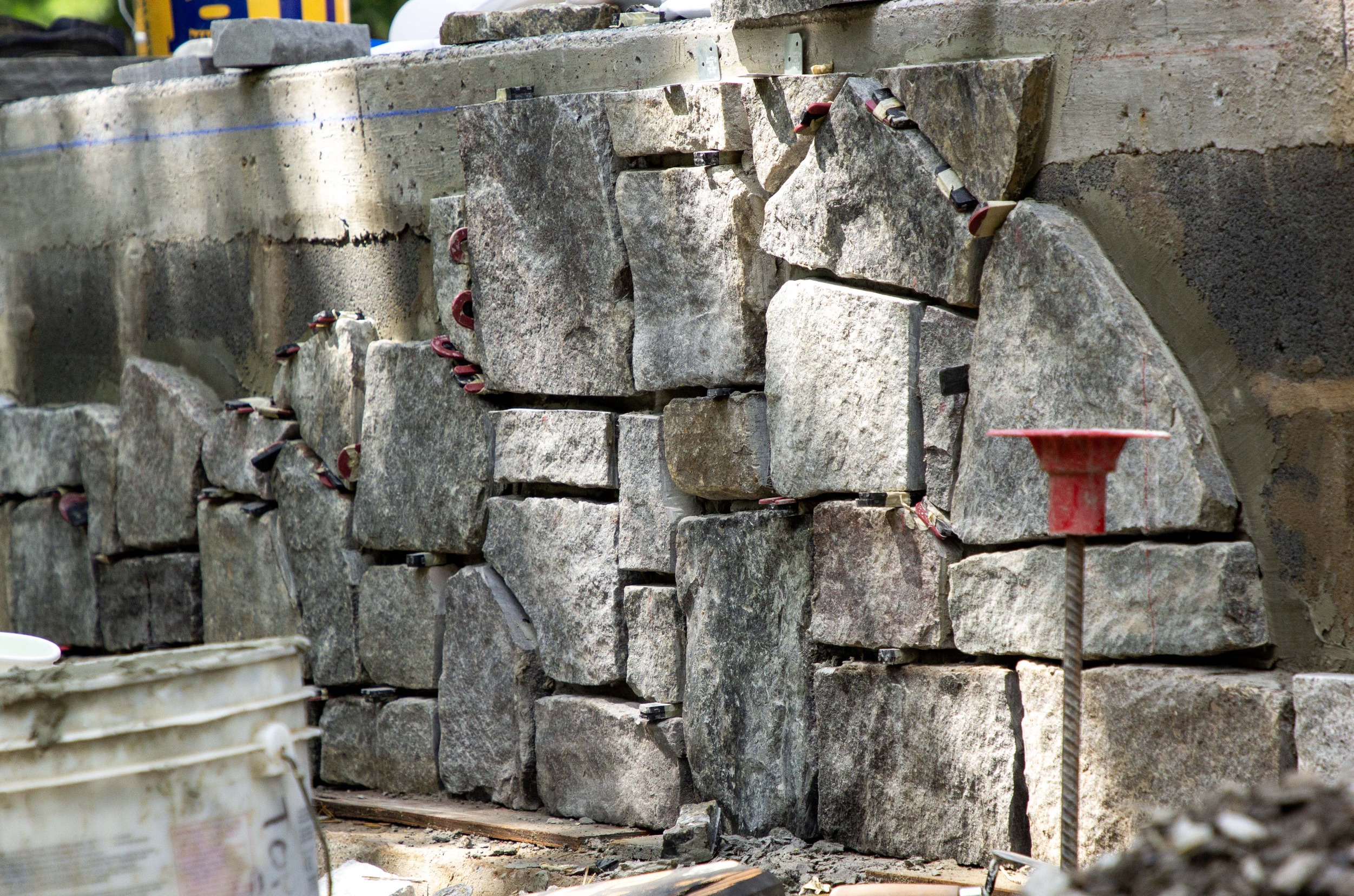 A stone veneer is being applied to a concrete block retaining wall. A white bucket and yellow bucket are also partially visible.