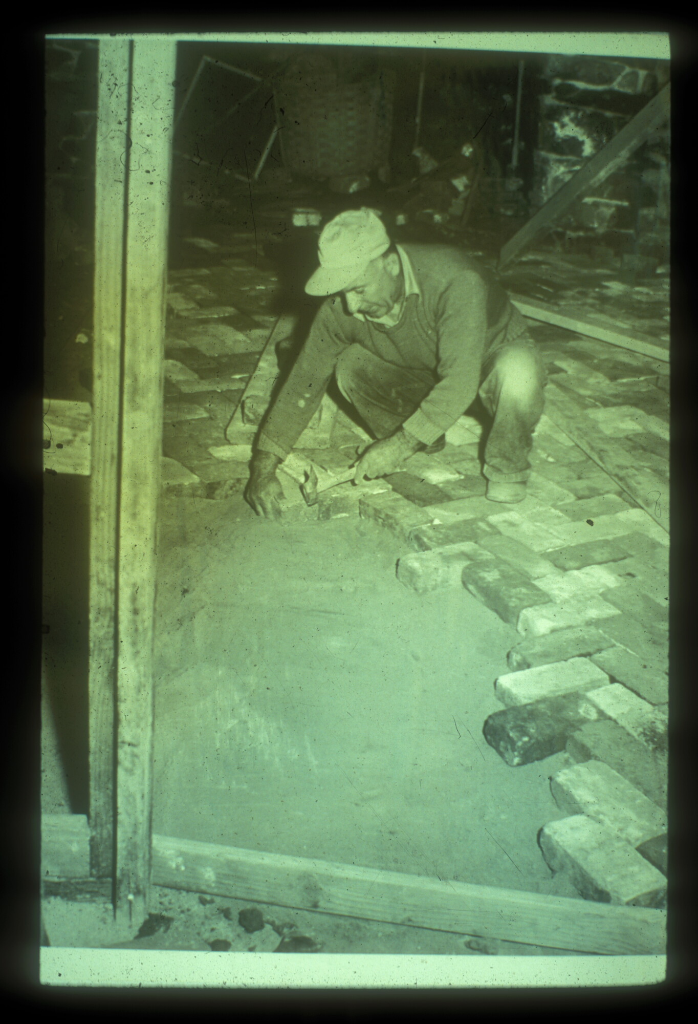 A man in a long sleeved shirt crouches down on a brick floor, tapping additional bricks into place with a hammer.