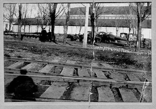 Railroad tracks in the foreground of the photo, a shadow of a man over the tracks.  Cannons stacks in the middle ground of the photo in between trees.  In the background is a building. 