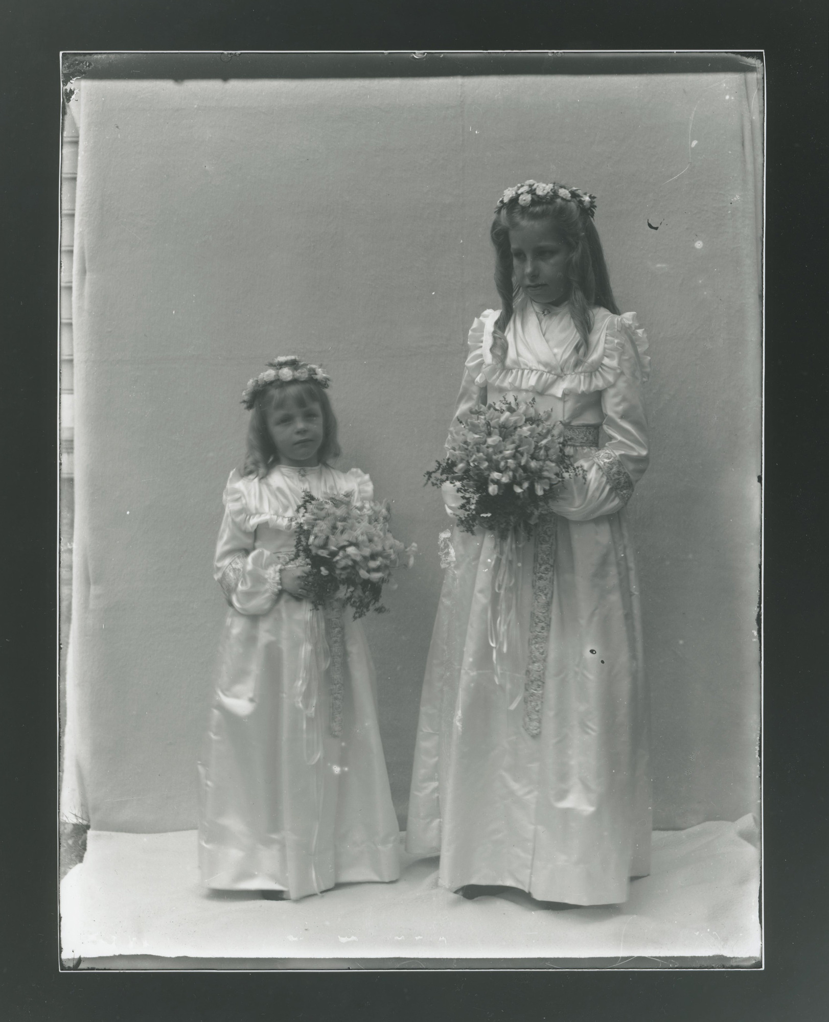 Two white children in ruffled white dresses hold hands. In free hand they each hold a bouquet of flowers. Both wear flower crowns.