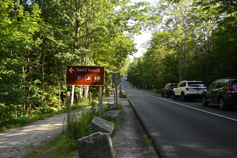 Roadway with cars parked on right side, and a brown 'Sand Beach' sign on left side pointing to entrance.