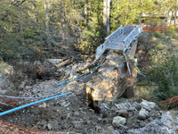 A blue boardwalk sits atop a thick concrete wall, both end abruptly with jagged edges. Behind the wall there are broken blue pipes, woody debris, and rock. There is orange safety fencing in the lower left corner and in the distance behind the boardwalk.