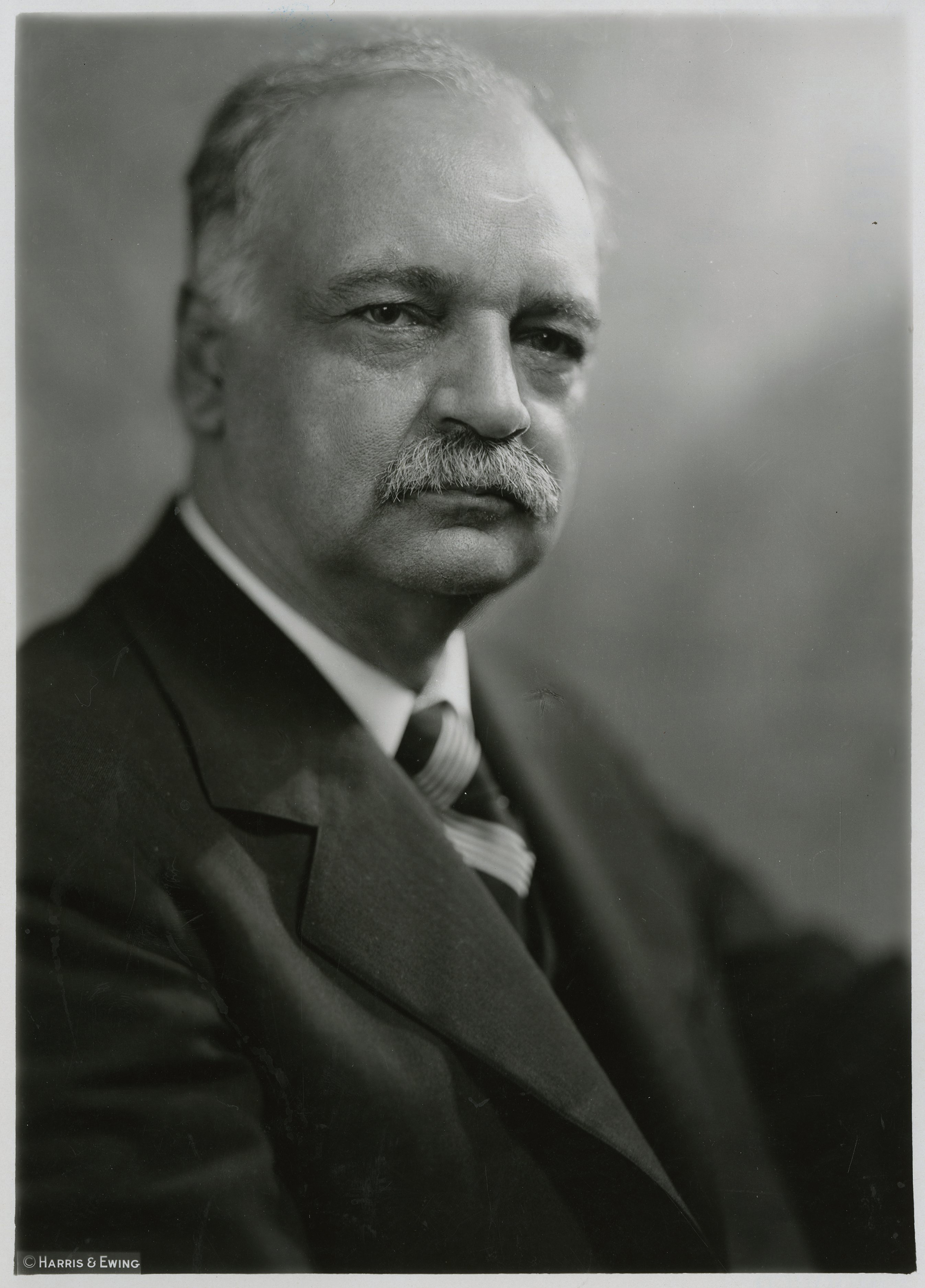 A 1928 bust portrait photo shows a serious gray haired and mustached man in a suit. 