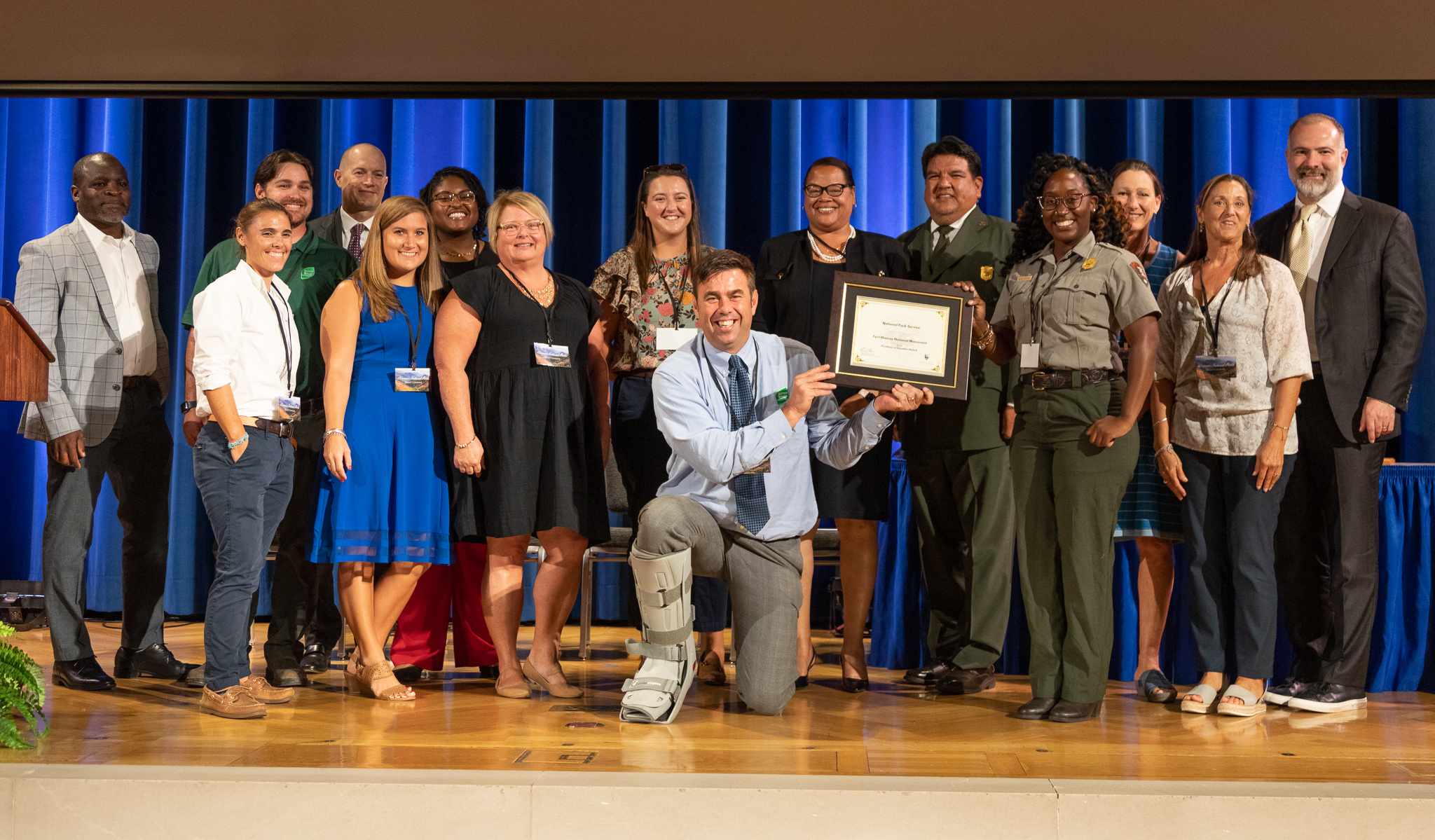 A group of fifteen smiling park rangers and staff accepts an award on a stage with the Director of the Park Service. 