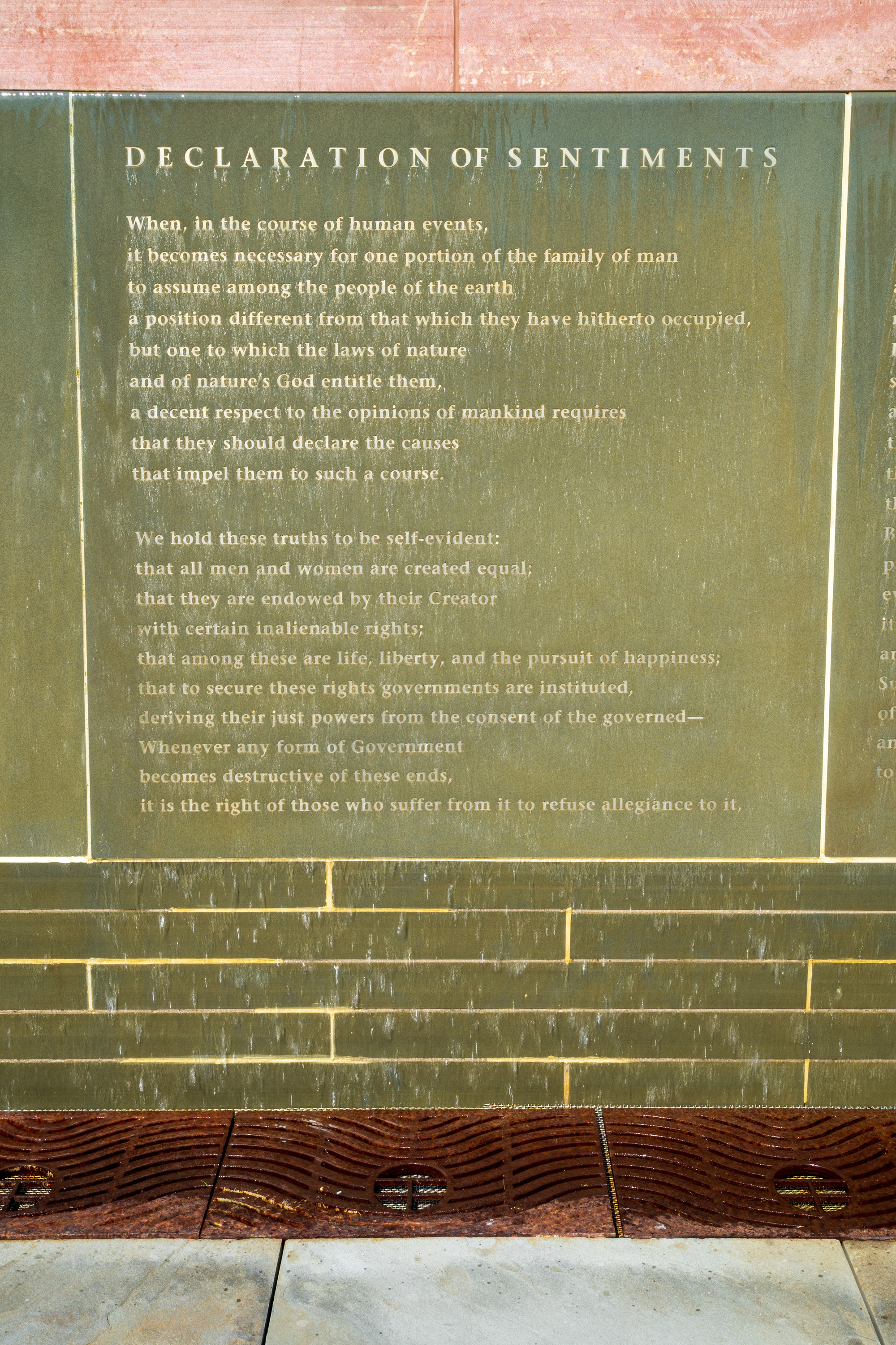 A greenish stone wall engraved with the first two stanzas of the Declaration of Sentiments, with water running over it.
