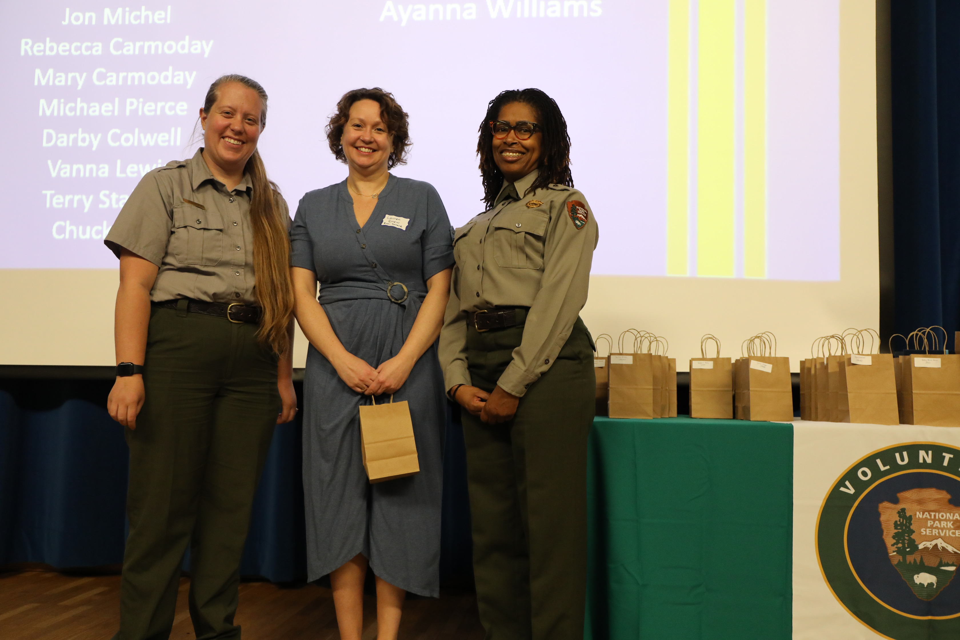 Three women stand side-by-side on a stage, smiling at the camera. On the left is a fair-skinned woman with light brown hair pulled back, wearing a short-sleeved gray uniform shirt and dark green pants. In the middle is a fair-skinned woman with short, curly brown hair, wearing a knee-length blue dress cinched at the waist, and holding a small brown paper bag. On the right is a dark-skinned woman with dreadlocks and glasses, wearing a short-sleeved gray uniform shirt and dark green pants. Behind them, a projector screen displays names, and to the far right, a table with a green tablecloth holds several brown paper bags. A banner with the "National Park Service Volunteer" logo is also visible on the table.