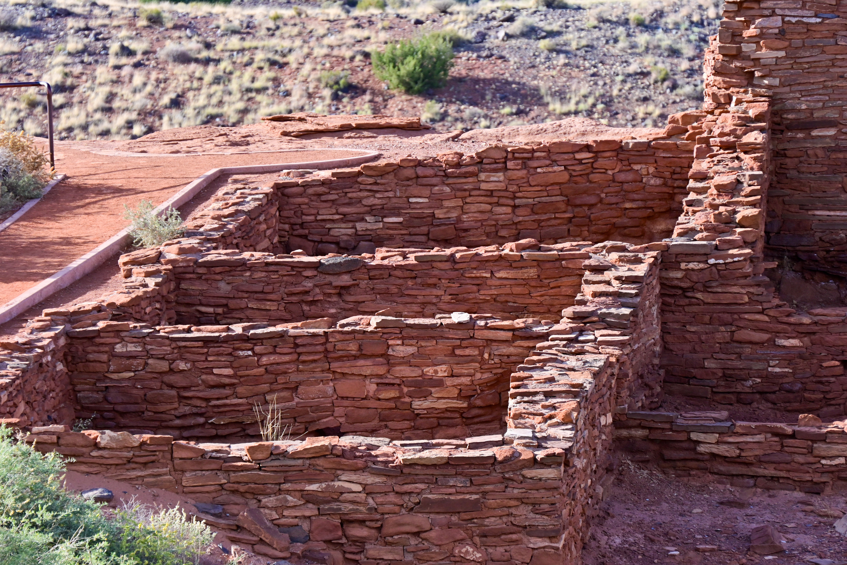 Walls of three distinct rooms formed of stacked red sandstone sit next to a red sand path.  