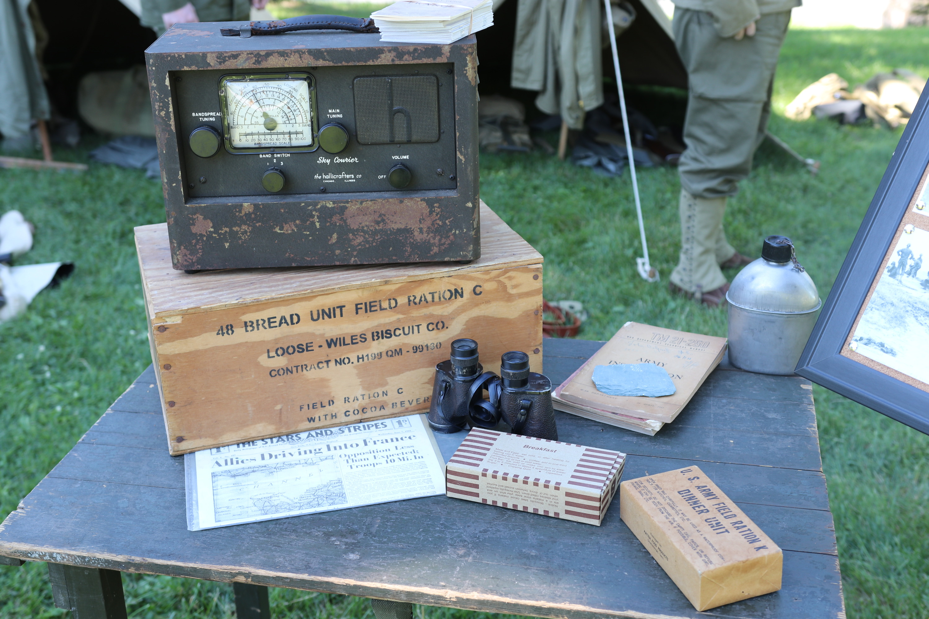 A WWII era radio sits on top of a rations box, with other soldier items on a table. 