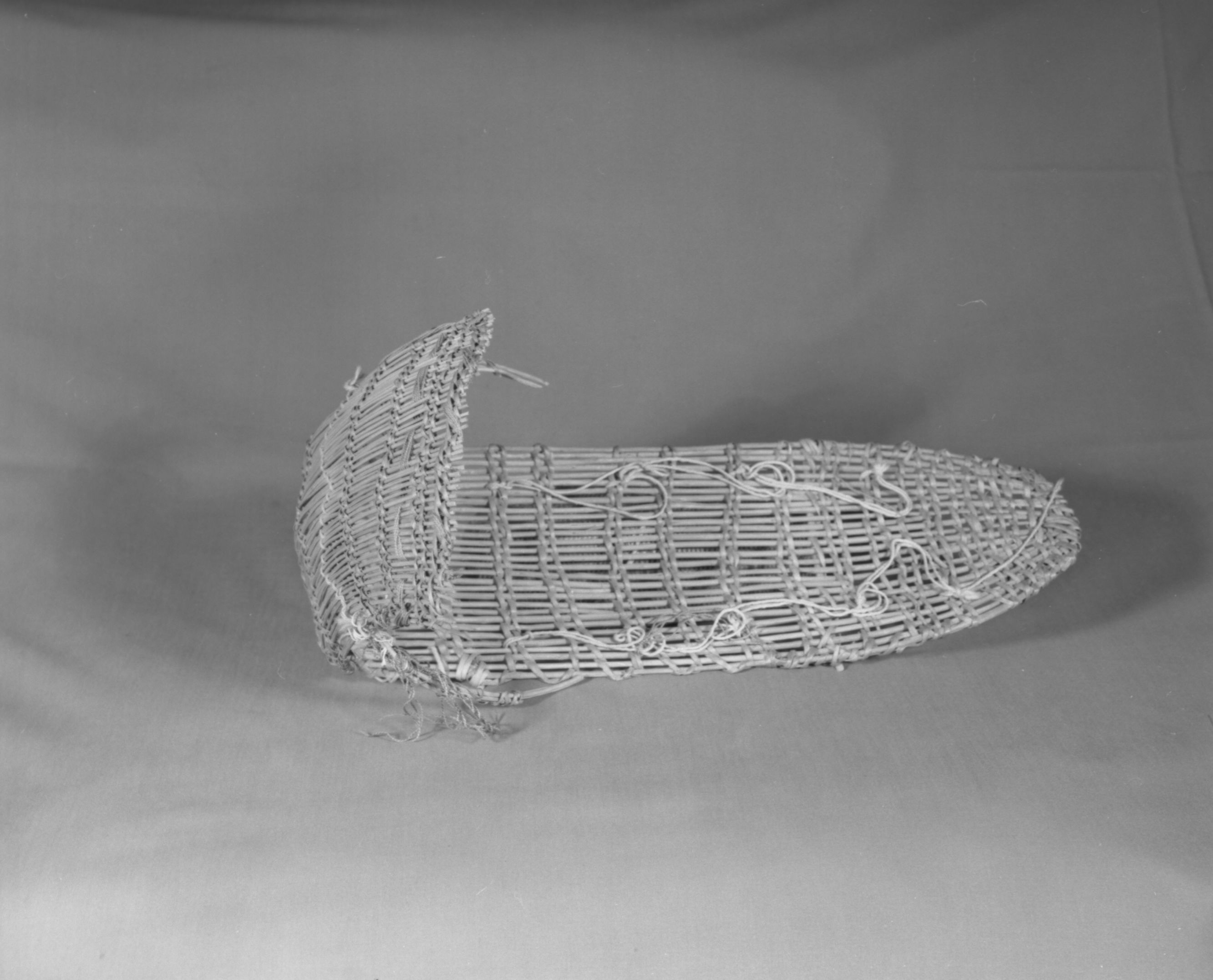 Private Collection. Object: Miwok cradle basket. Dimensions: Backboard - 17 x 6 1/4" Hood - 7 1/4 x 9". Materials: Split bullpine, ceanothus cuneatus, four ply "s" twist commercial cotton string, red commercial string, possibly added on later. Technology: Twined up to right slant of turns, start is begun by bending warp rods over another rod. Rod bent around lower part of basket. Two additional strengthening rods also attached across reverse of basket. Hood has one row of three strand twining near lower edge. Collection History: Made for the owner's doll, when she was a young girl, by Matilda, a Sonora Miwok, c. 1920. Photographed by Michael Dixon. Catalogued by Craig Bates and Barbara Beroza. Yosemite Collections, National Park Service.