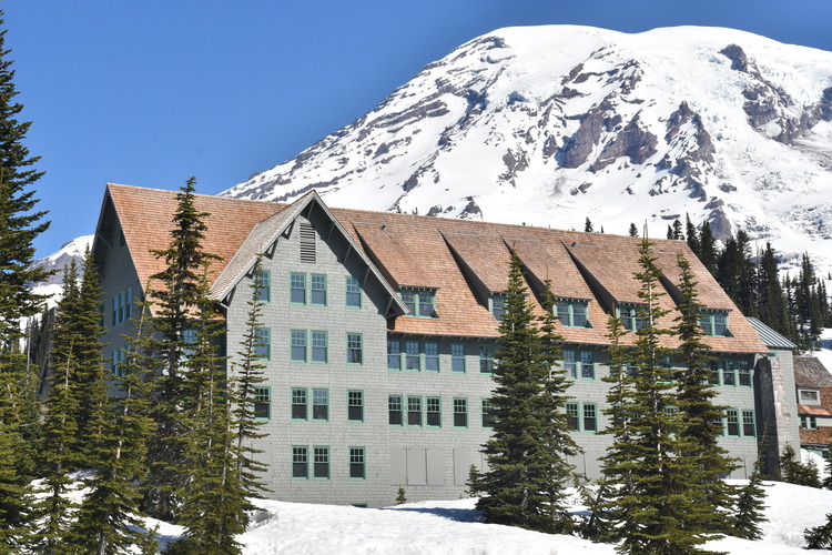 A four-story wood building with grey shingled sides and an angled roof in front of a glaciated mountain. 