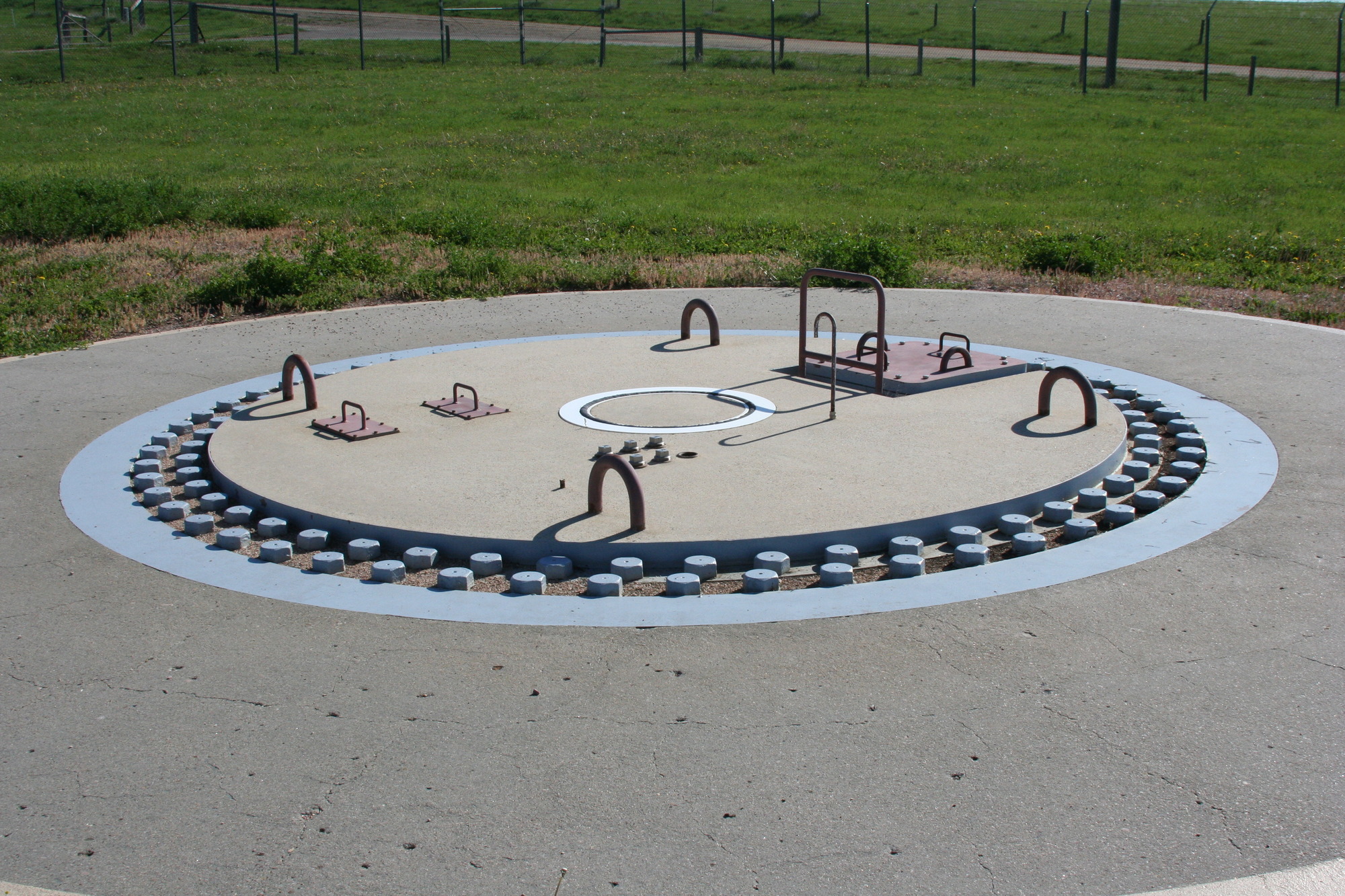 In a field of grass, a large concrete circular pad with a yellow stripe around the outside and a blue circle in the middle. Large red metal bars come out of the ground inside it like giant handlebars. 