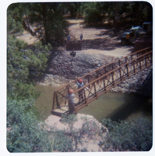 Two men guiding new Grotto footbridge into place as it is lowered on pulley system. Note bank revetments in background.