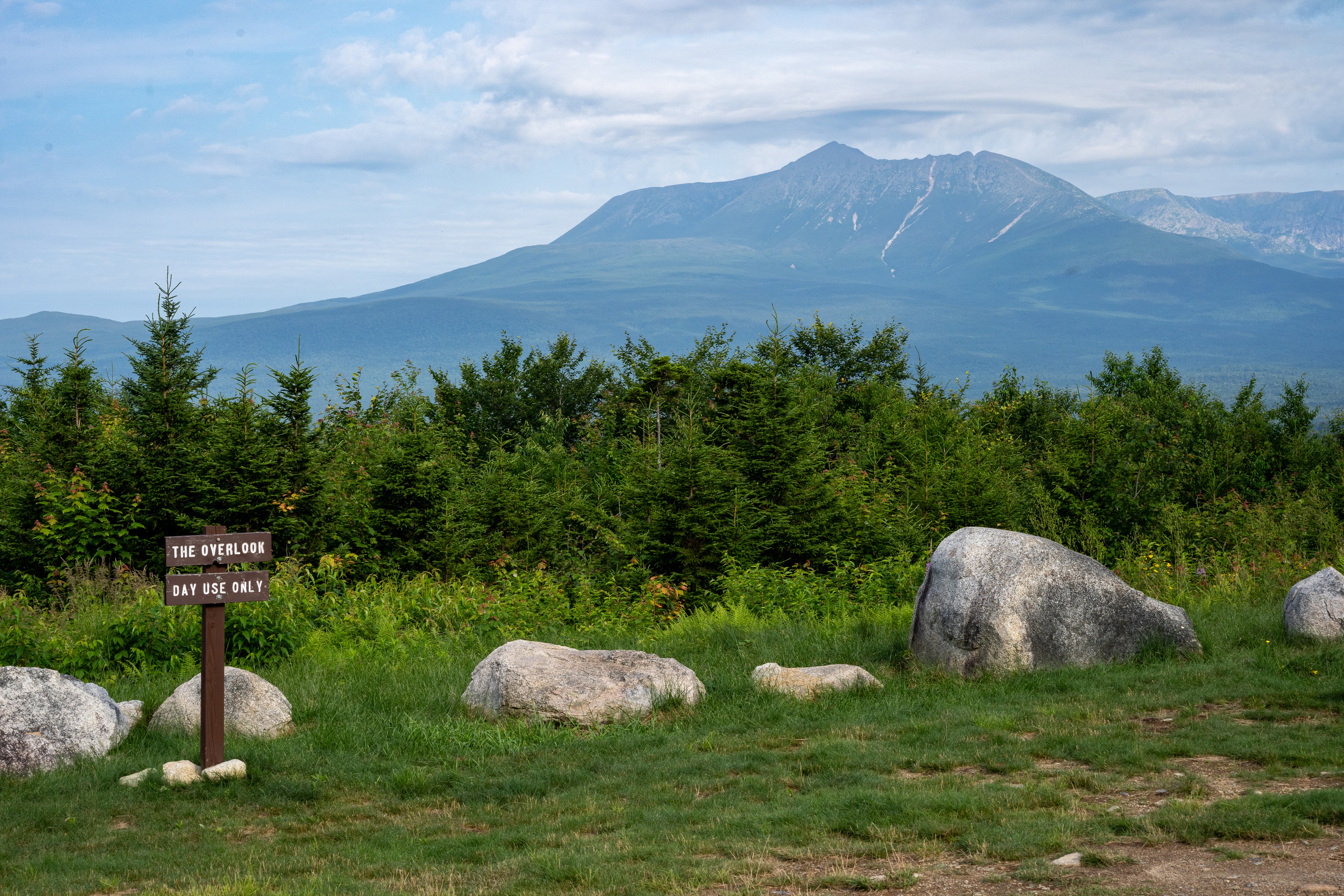 A mountain against a cloudy blue sky. Boulders and trees are in the foreground. A sign reads "The Overlook - Day Use Only"