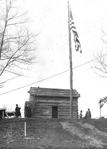 A black-and-white image shows an American flag rising above a small log cabin. 