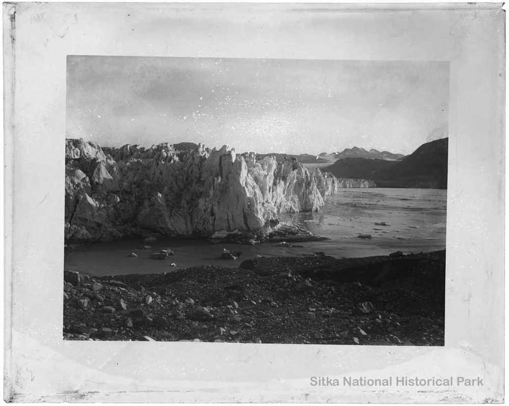 A positive of a tidewater glacier with a gravelly shoreline in the foreground and mountains in the background.