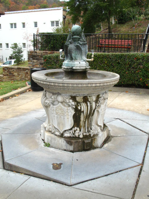 An elaborate marble fountain base with a shallow round basin topped with bronze eagles, flags, standards, and shields. 