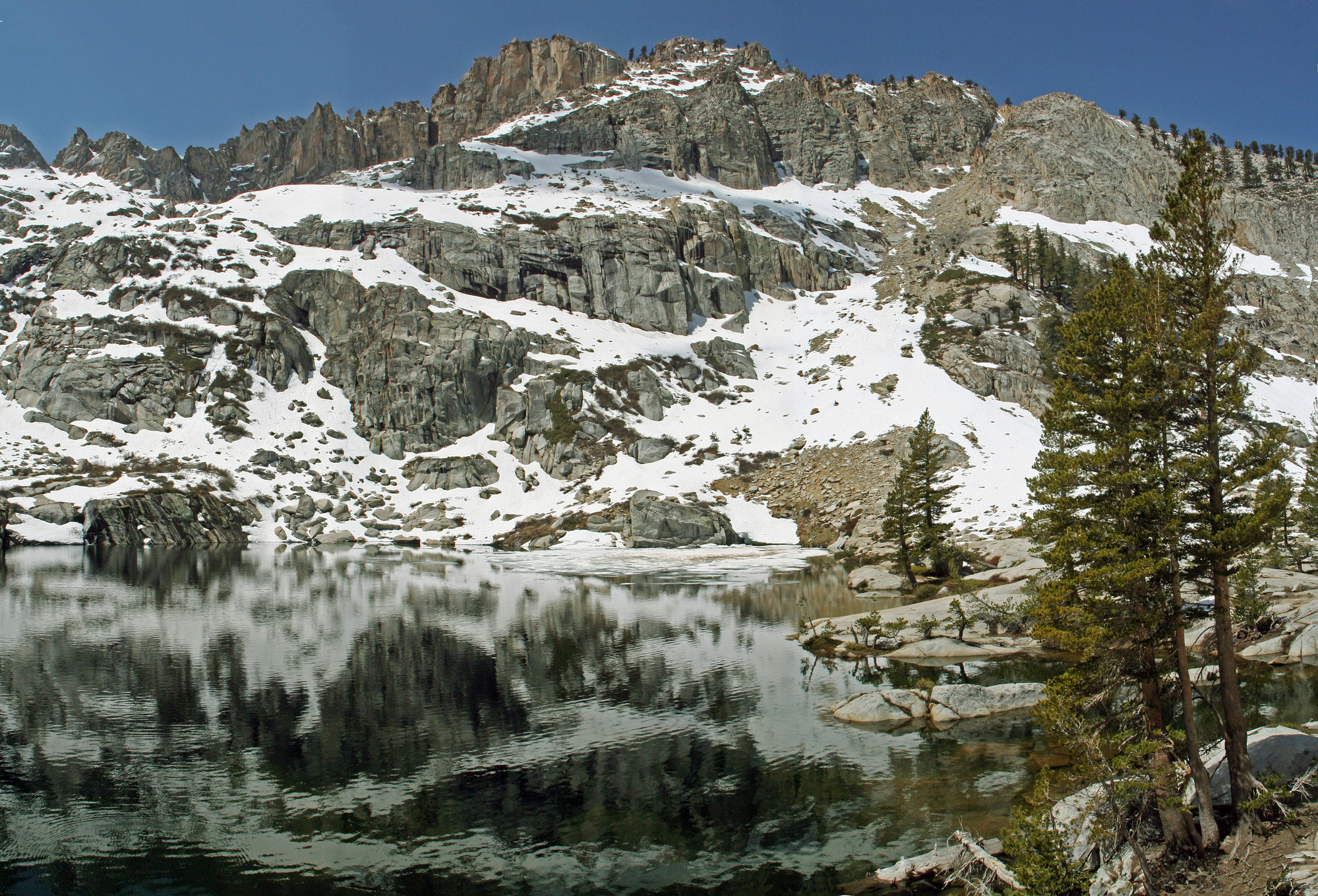 Emerald lake in front of a snowy and rocky mountain. There are a few trees by the lake.