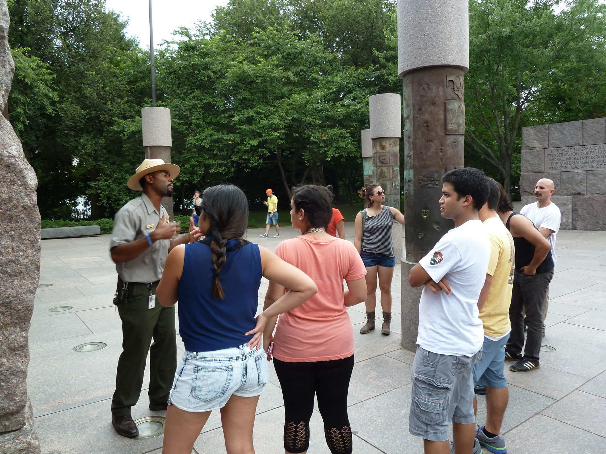 A park ranger talks to a group of 7 people as they stand around several metal poles with designs on them. 