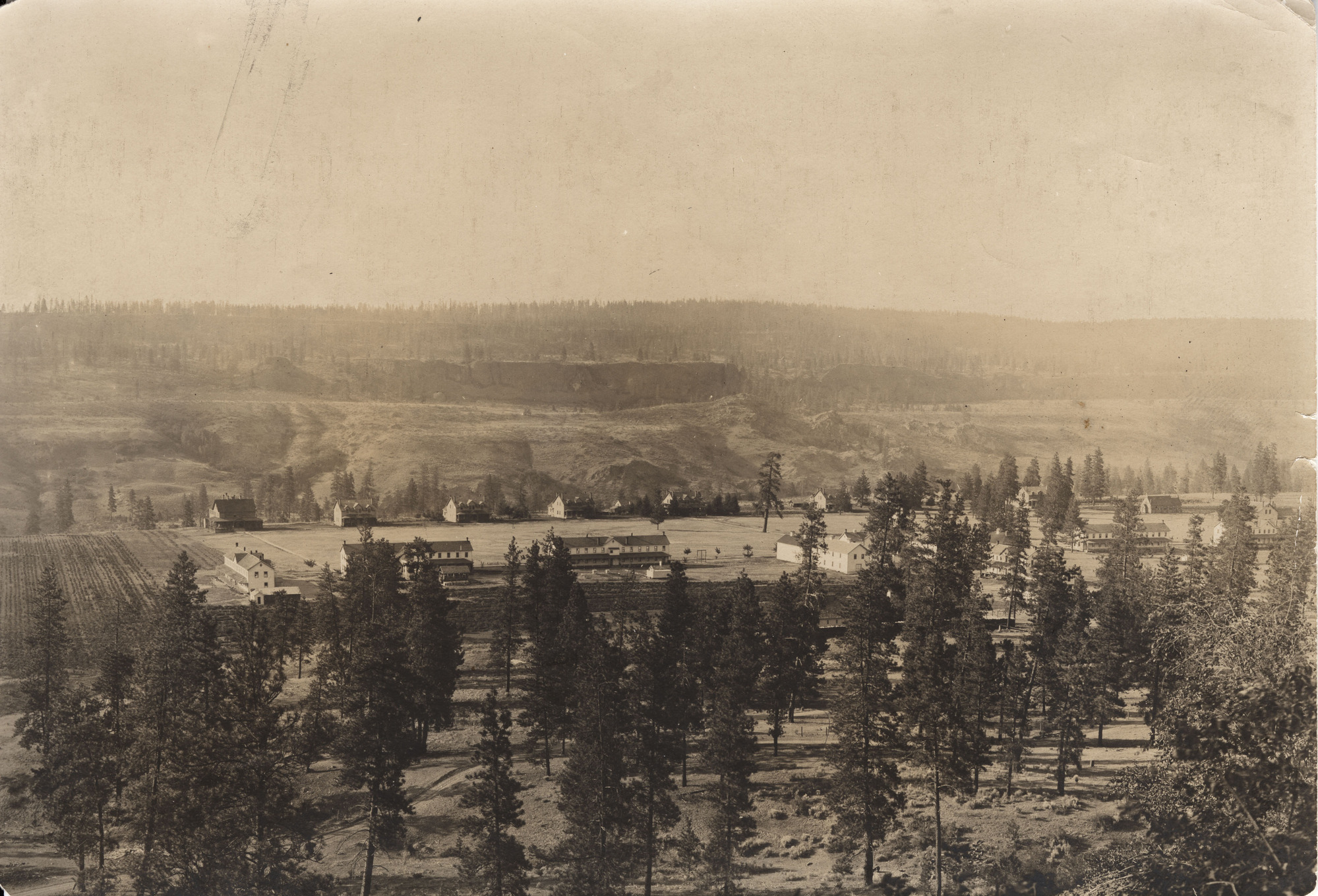 Sepia toned photograph of a complex of buildings in a wide forested valley