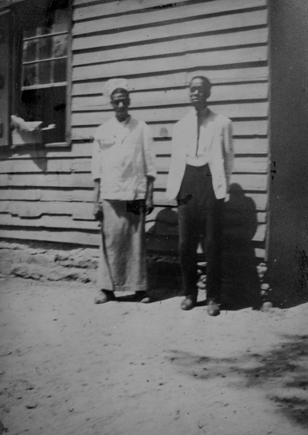 Black and white image of a man and a woman standing in front of a wooden structure wearing the work uniforms of domestic servants in the early 20th century.