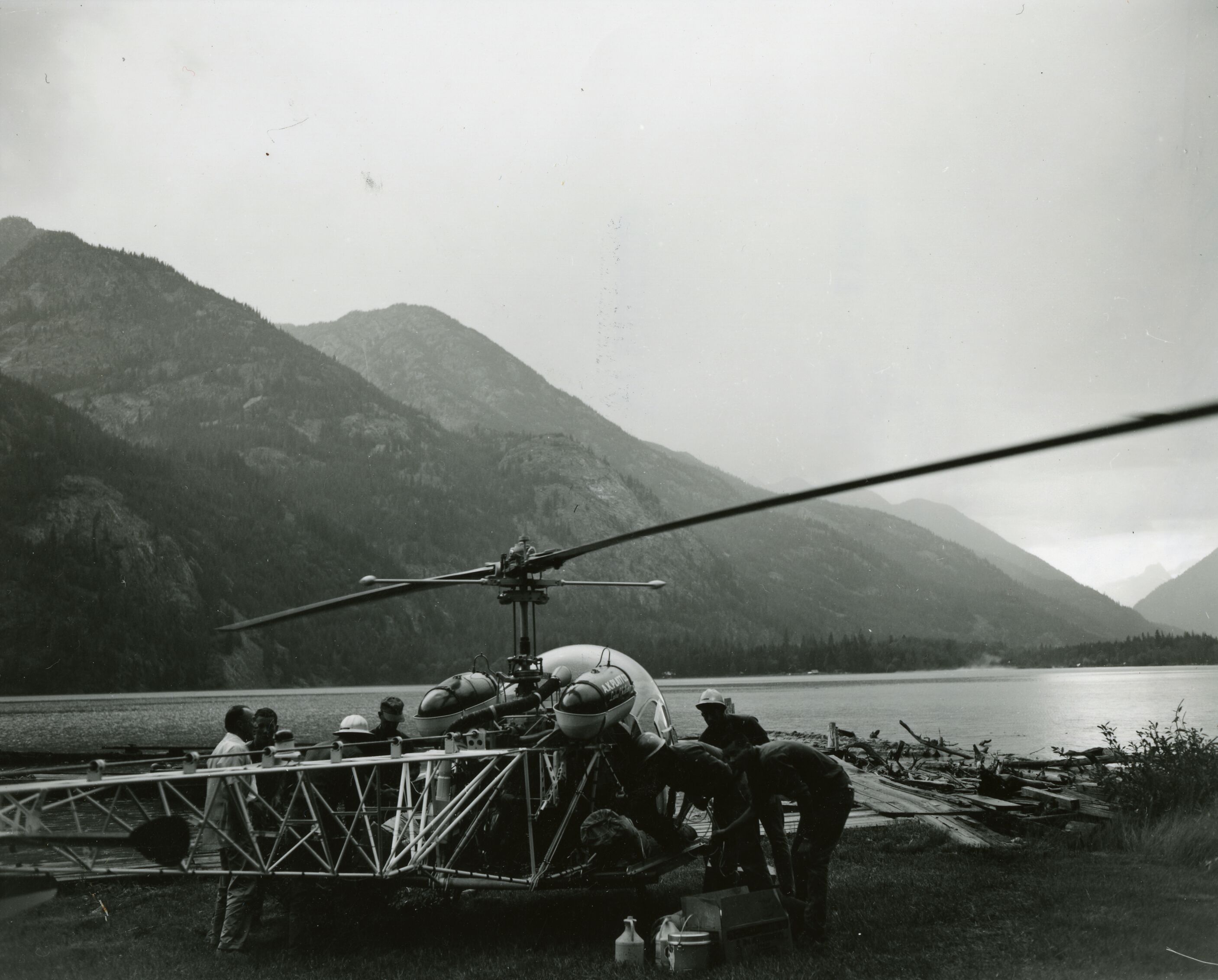 Several people crowded around a helicopter.  A lake and low mountain ridges are in the background.
