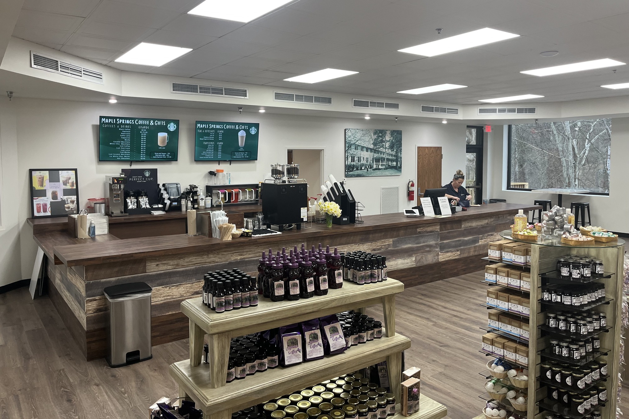 A woman stands at a long wooden counter with equipment used to make coffee and drinks. Large menu screens and paintings hang on the wall behind the counter.