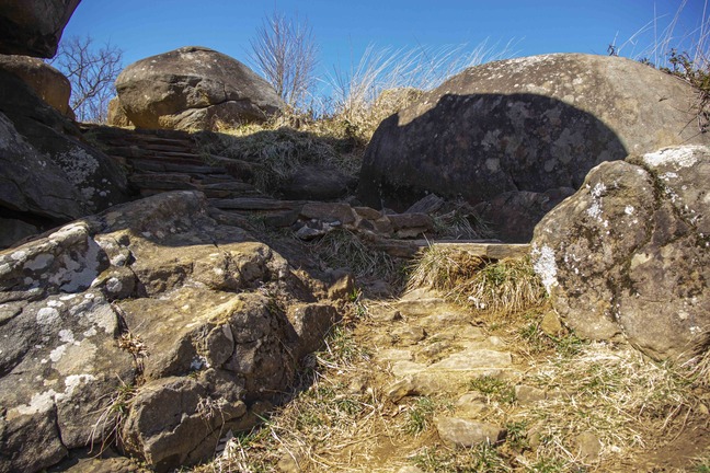 There are narrow, jagged stone steps leading up the side of a small slope and to the left. On either side of the steps are large boulders. Some of the boulders are casting their shadows over the top of the steps. There are bare treetops visible in the distance beyond the top of the slope.