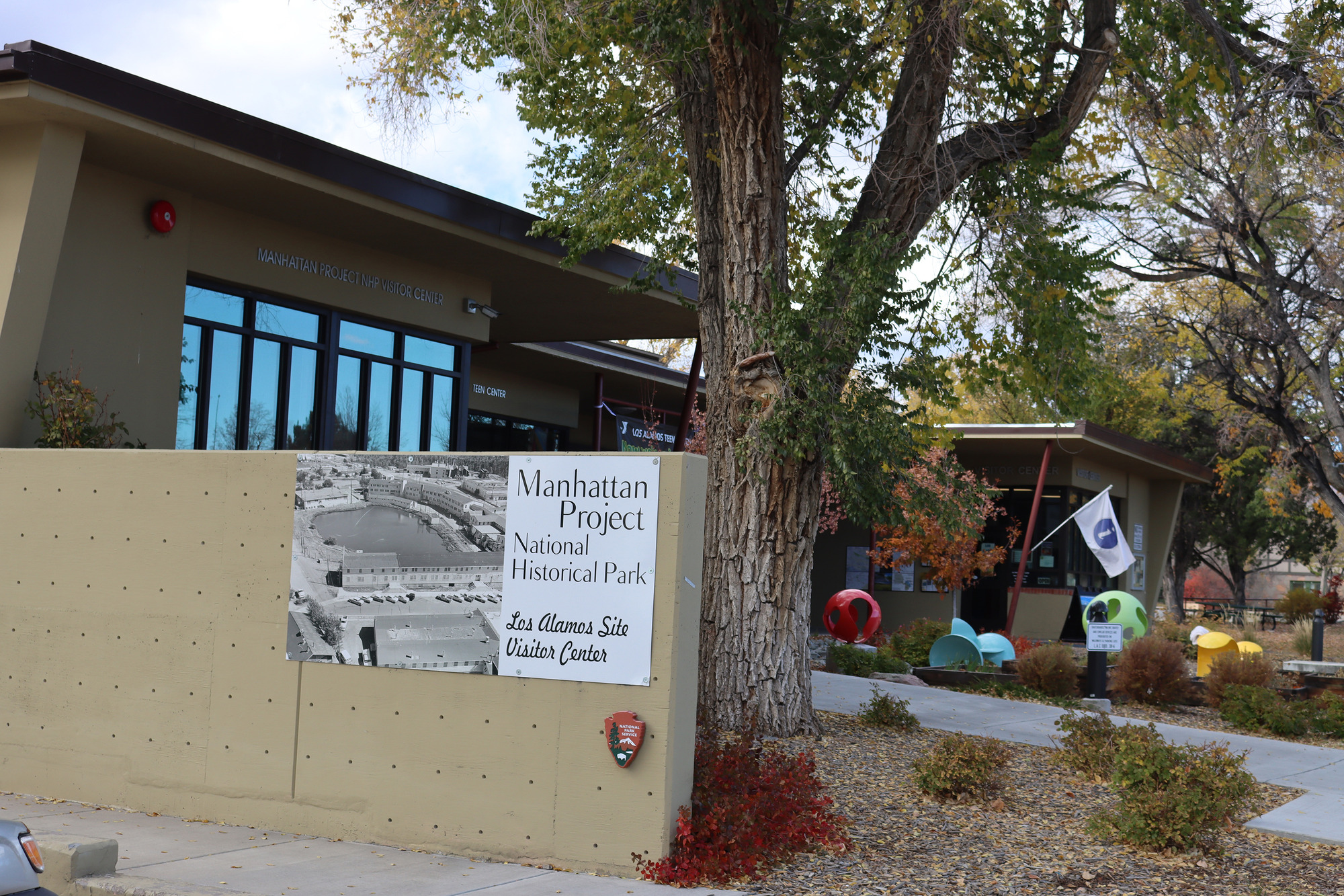 A tan single-story building with a black and white sign that reads, "Manhattan Project National Historical Park: Los Alamos Site Visitor Center.” In front, a small manicured courtyard with a large tree and a white flag with a blue “i” signaling information available here. 
