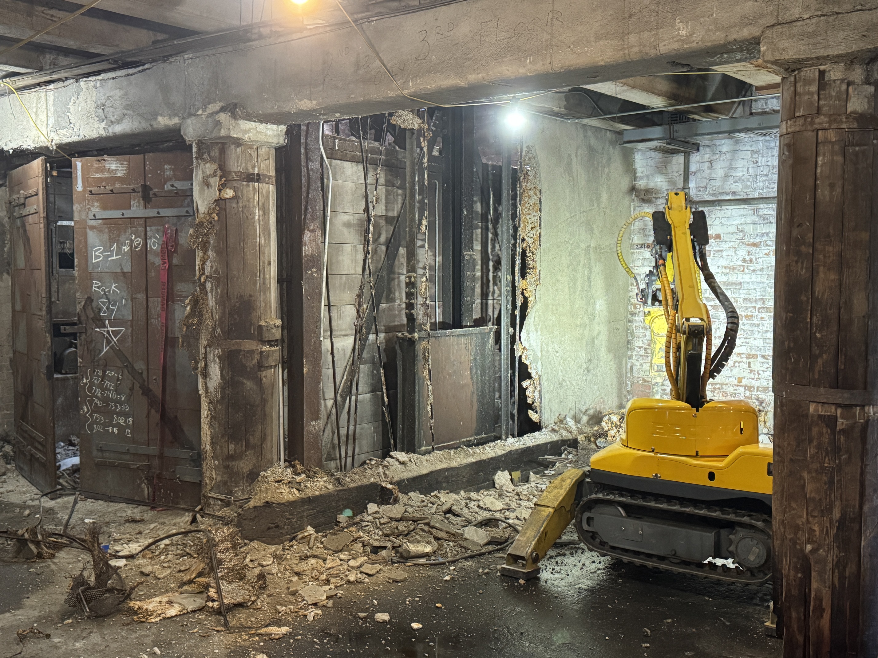 A yellow robot with a crane-like arm in a room in an industrial building surrounded by debris. 