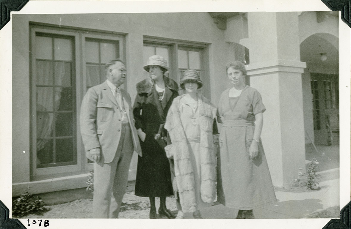 This is an historic black and white photograph from the Scotty's Castle Historic Photograph Collection, Death Valley National Park of four people in front of a large Spanish style house. Two women in middle in cloche hats, dresses and fur-trimmed coats. Man on left in suit. Woman on right in dress. Number in black ink in lower left corner.