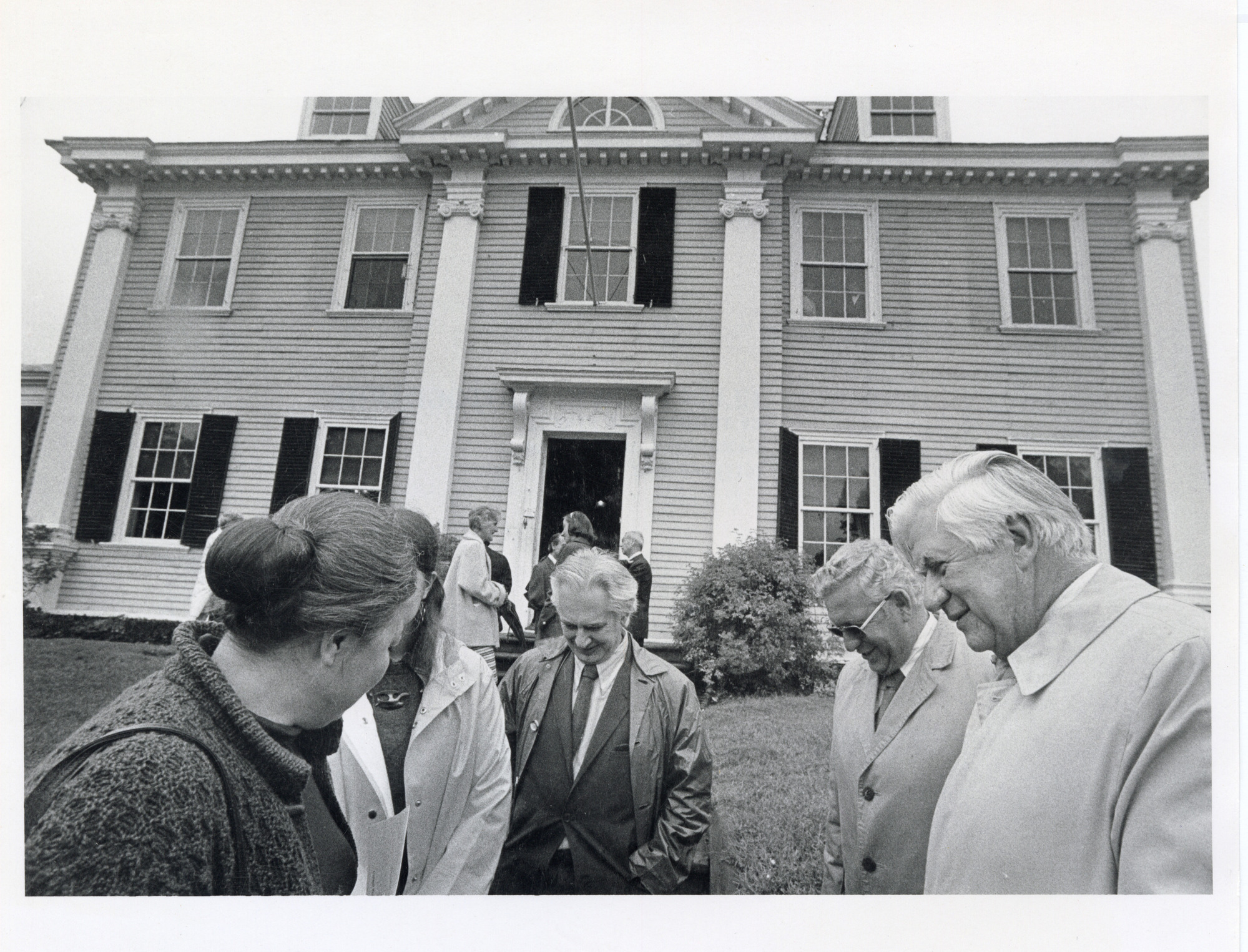 Black and white photograph of group of people gathered on front lawn of large mansion with some missing shutters and open front door.