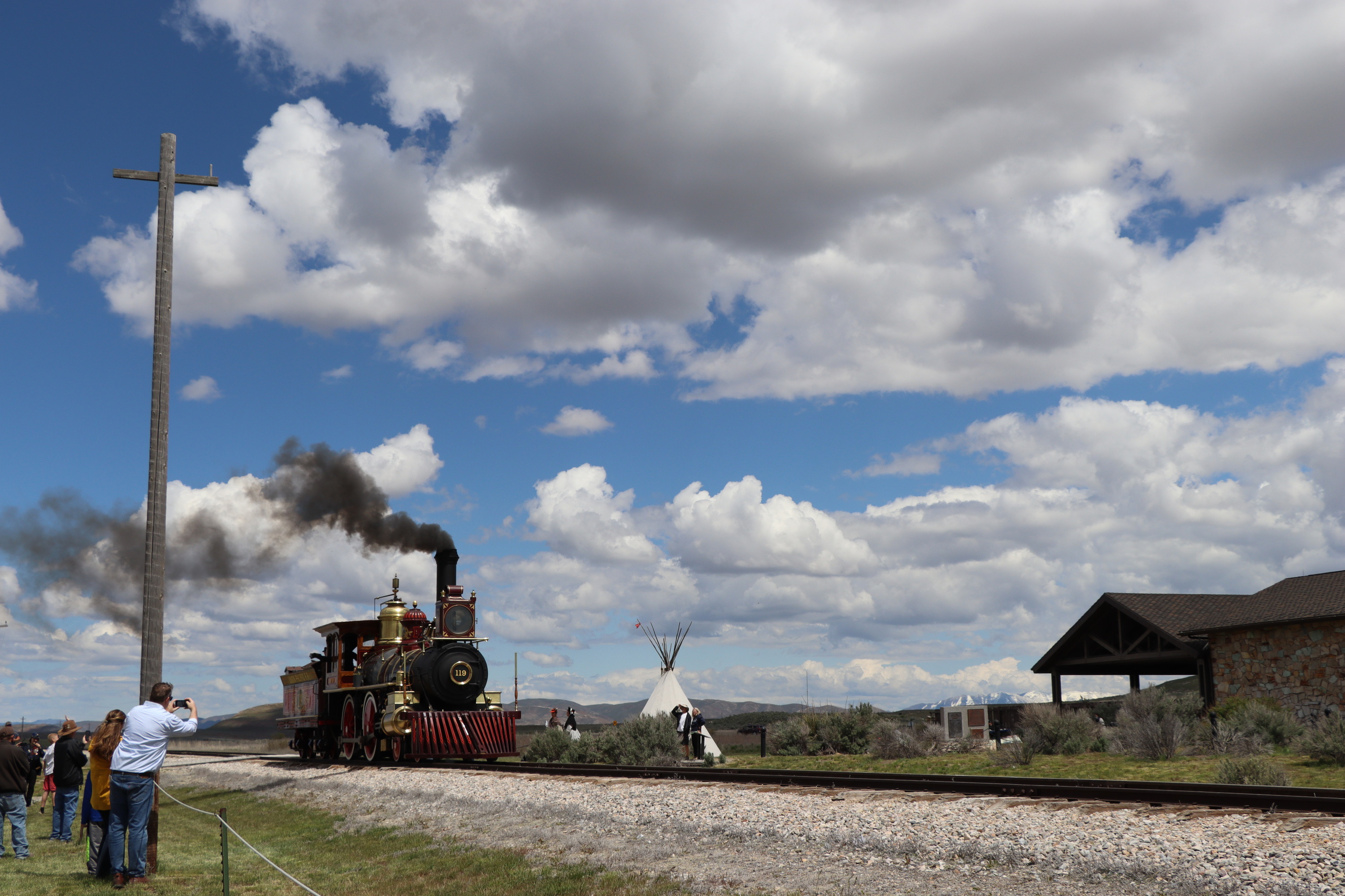 The red, black, and gold steam locomotive No. 119 moves down the railroad track while people watch. It releases black coal smoke against the blue sky. A teepee, building, and telephone pole are visible.
