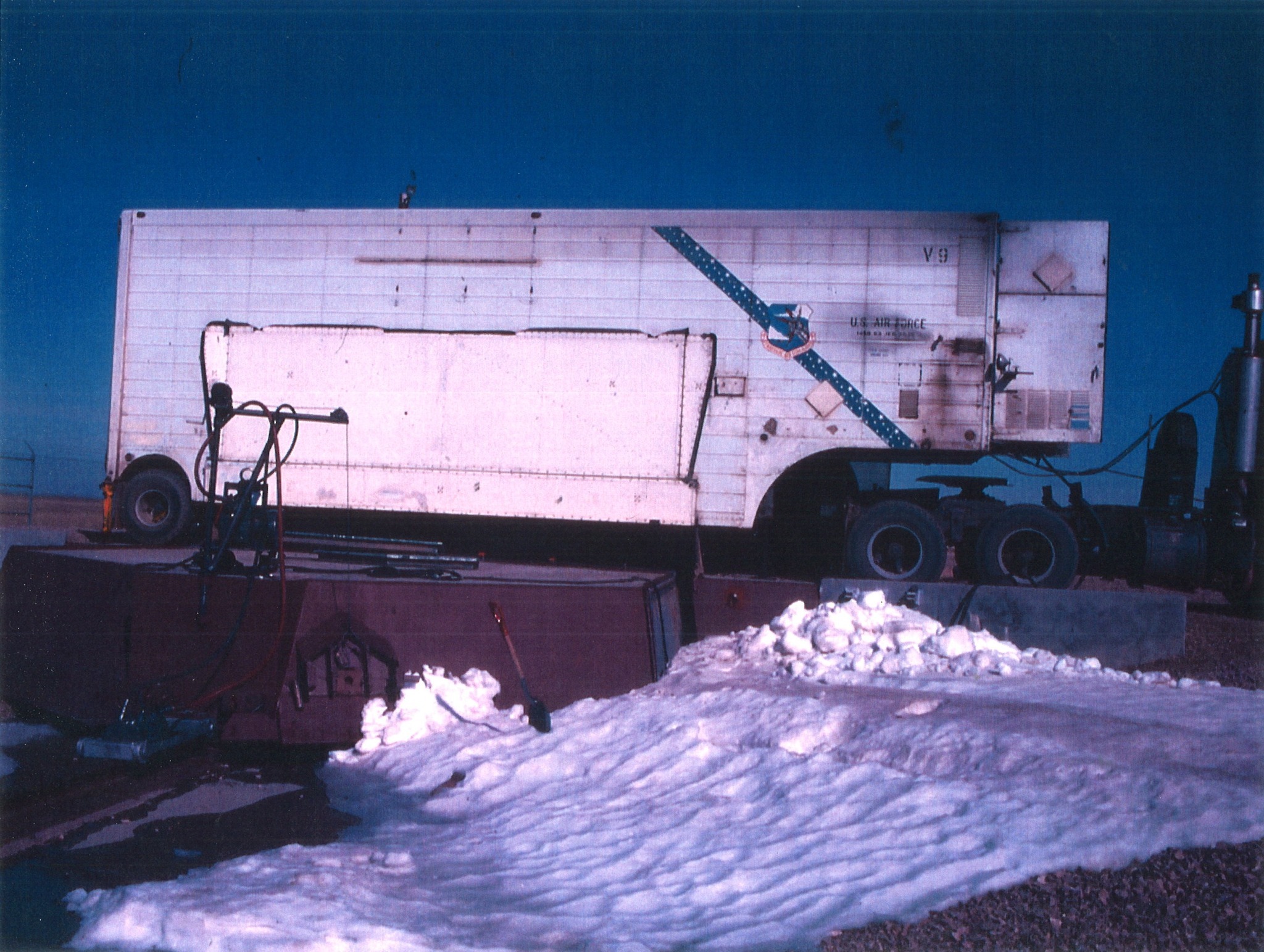 A long white truck parked across a concrete pad in a field of snow and gravel.