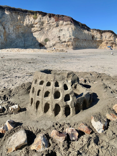A small sand sculpture of the Coliseum in Rome, Italy. The sculpture is a bit more than a foot tall, is circular in shape with alcoves carved into the exterior walls, and part of the wall collapsed on the right.