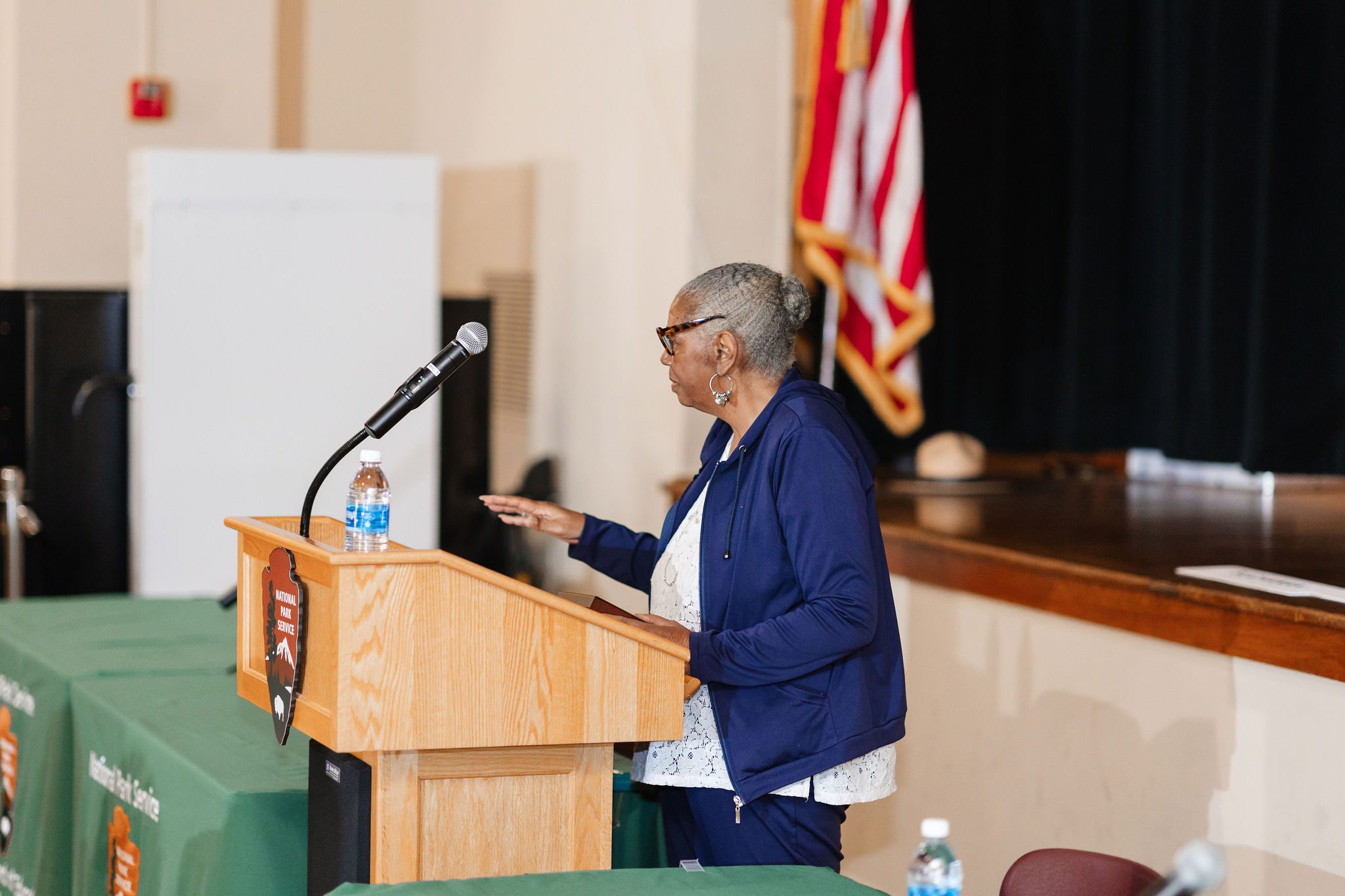 A shorter african american woman in a blue jacket speaks at a wooden lectern with the NPS arrowhead emblem on it. The lectern is in front of a wooden stage with the American flag on it.
