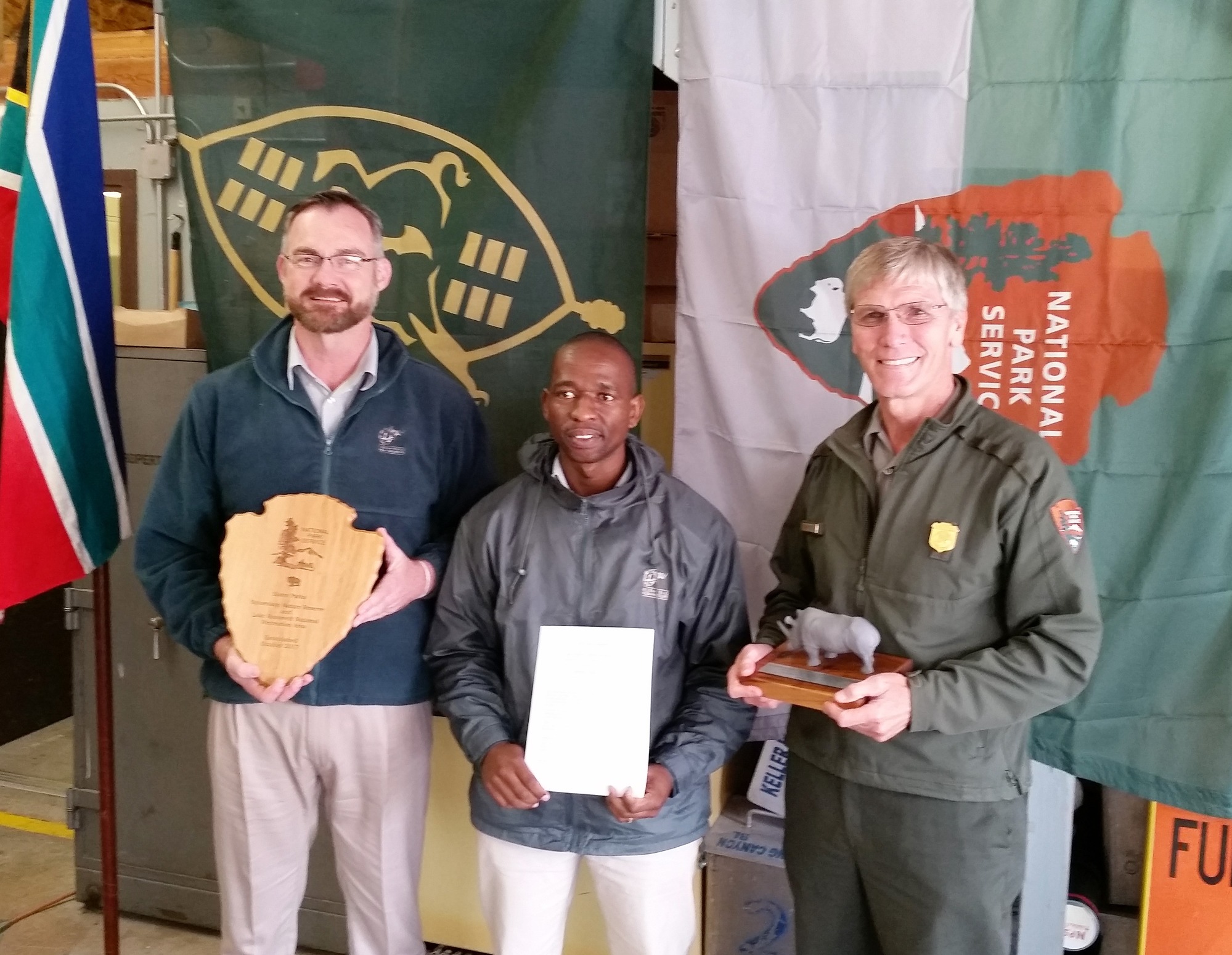Three men standing in front of the flags of the National Park Service and South Africa hold the sister park arrangement document, an NPS arrowhead and a rhino.