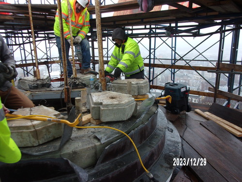Workers in safety and high visibility gear remove light grey marble stones from a circular assembly. Scaffolding surrounds the work area.
