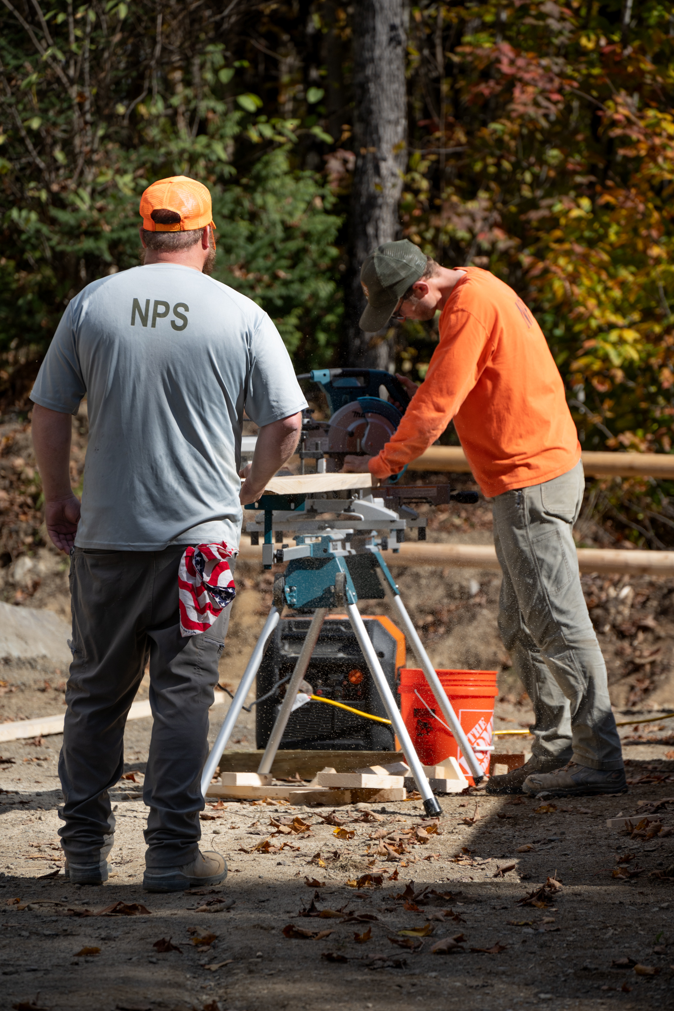 Two employees work on cutting lumber at a construction site. The background is forested with green, red, and yellow leaves. 