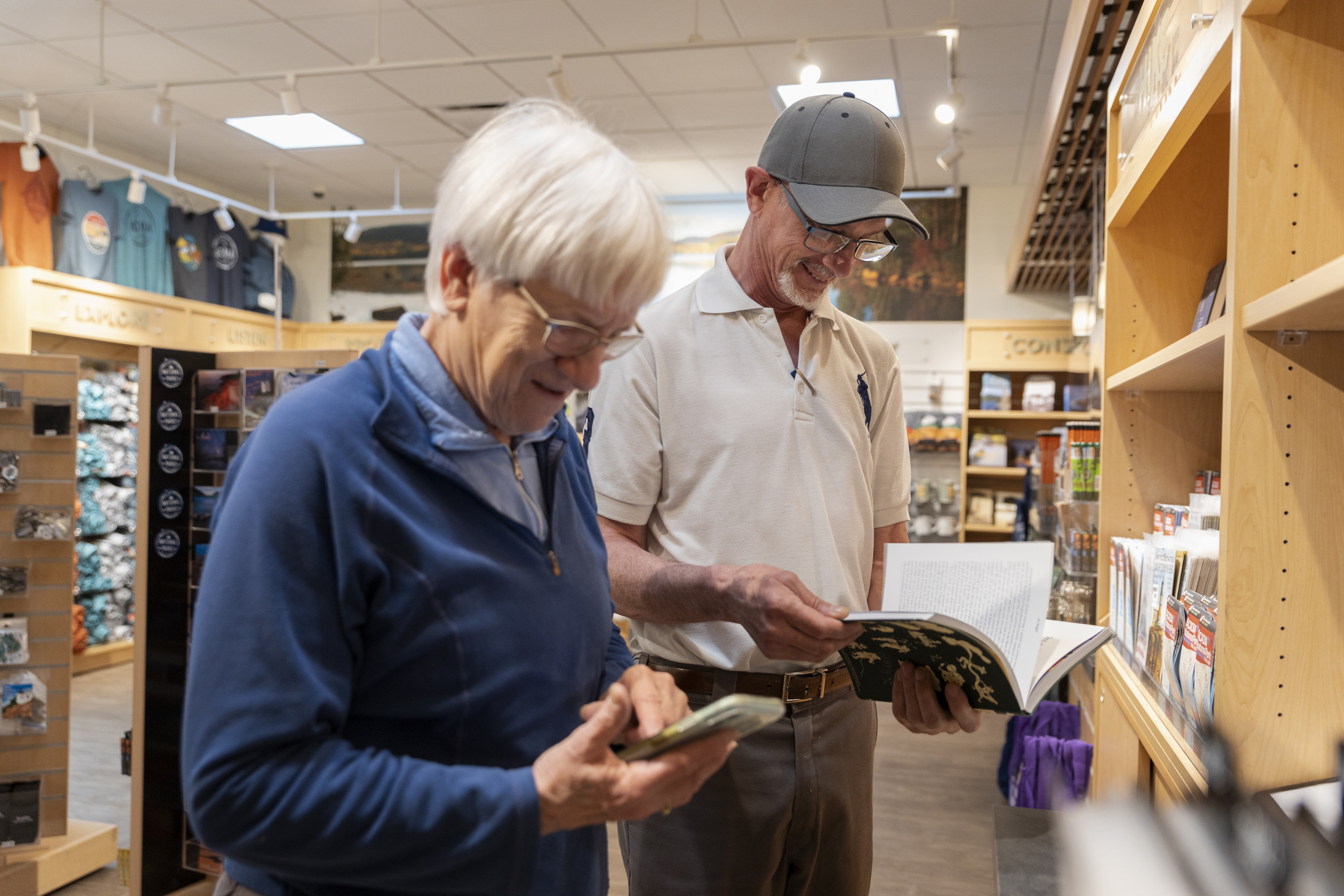  Two older adults exploring merchandise in Acadia's Park Store, with the woman examining her phone and the man reading a book, amid shelving filled with souvenirs.