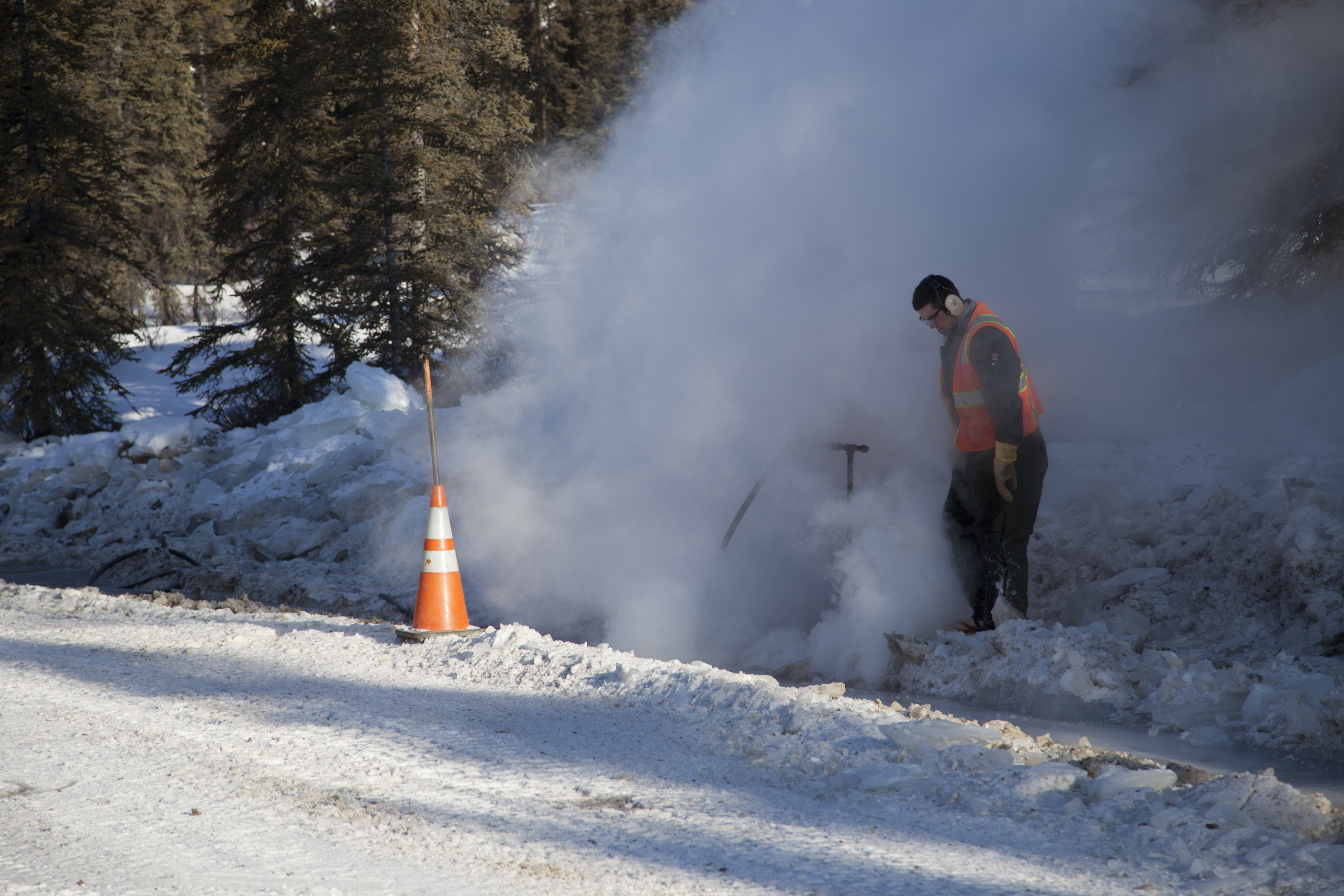 man standing by a roadside, covered in steam emerging from a culvert under the road