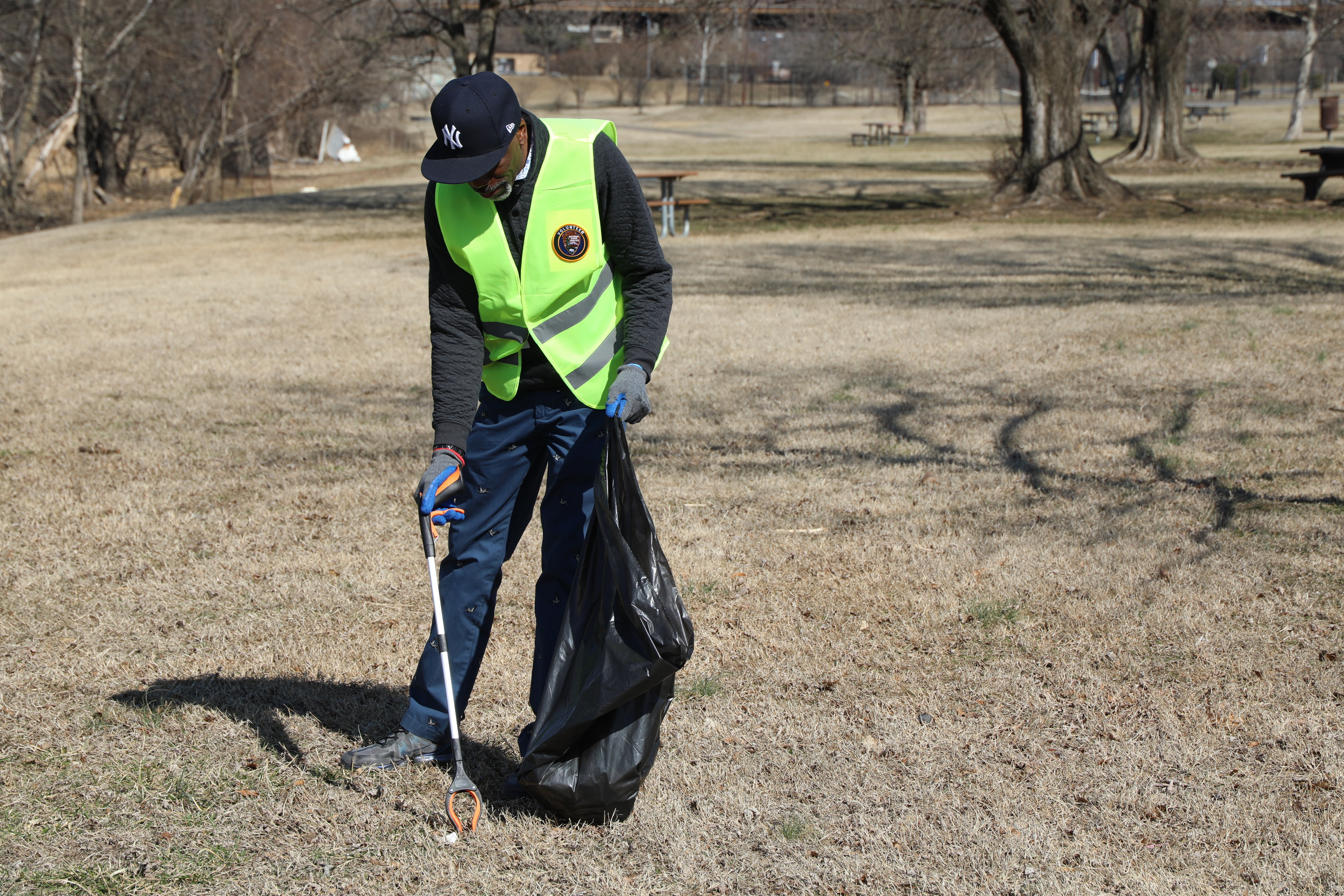A person wearing a dark long-sleeved shirt, dark blue pants, a bright yellow-green reflective vest, and a dark baseball cap with a white "NY" logo is standing in a large, grassy park. They are holding a long white trash picker in their right hand, aimed at the ground, and a black garbage bag in their left hand, which is open. The grass is dry and light brown. In the background, there are bare trees, picnic tables, and other park features under a clear, sunny sky.