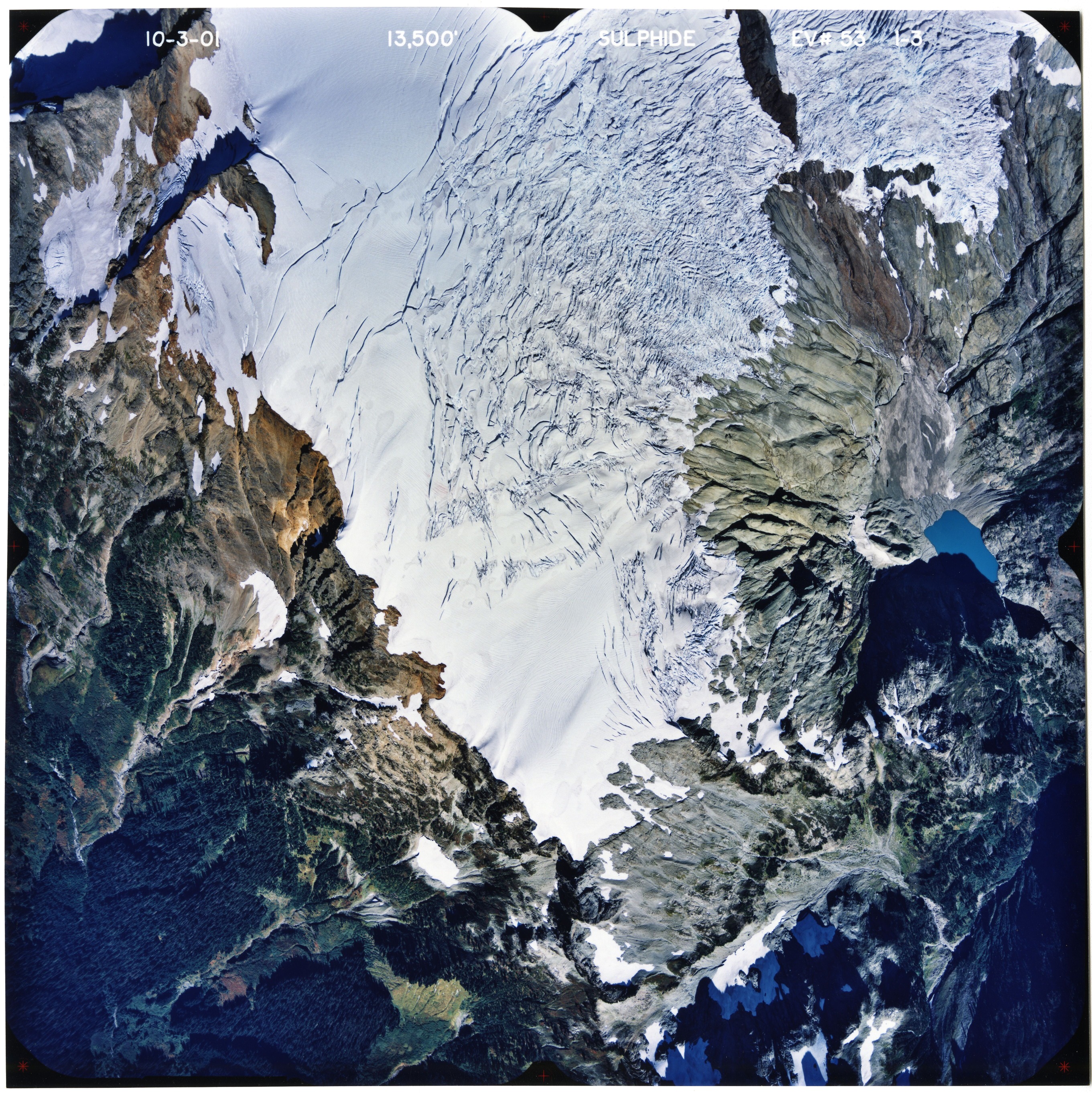 Color aerial photograph of glacier on mountain peak with forested valleys below.
