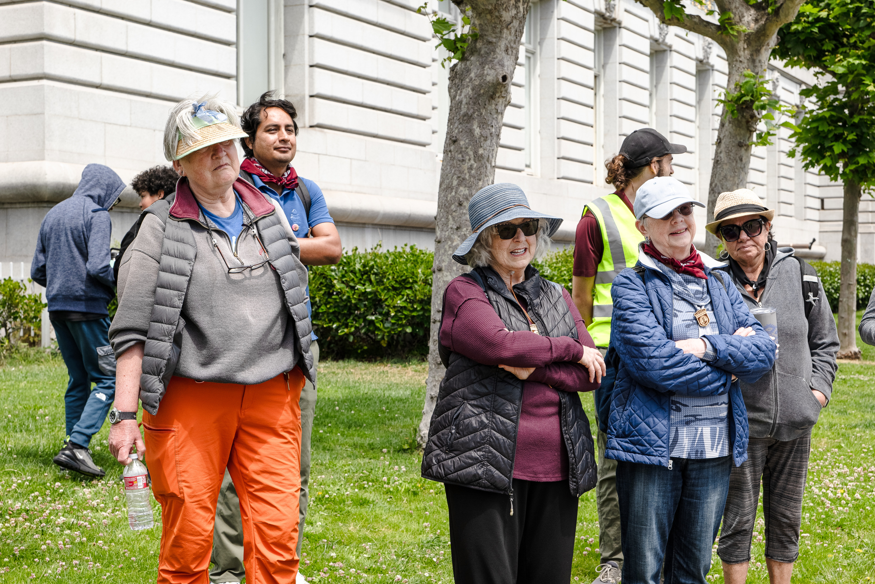 A group of people wearing jackets, bandanas, hats, and sunglasses gather on a grassy lawn with trees