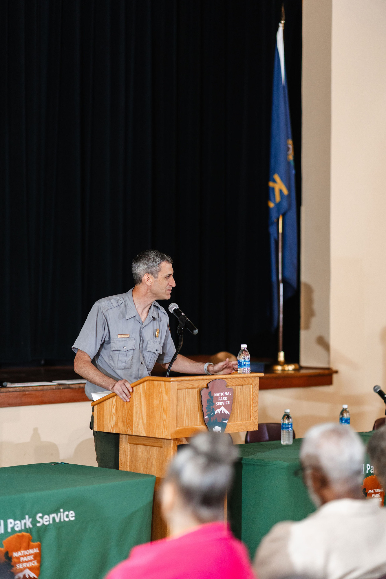 A tall white park ranger with short dark hair stands behind a wooden lectern speaking into a microphone. The lectern is in front of a wooden stage with an American flag resting on it.