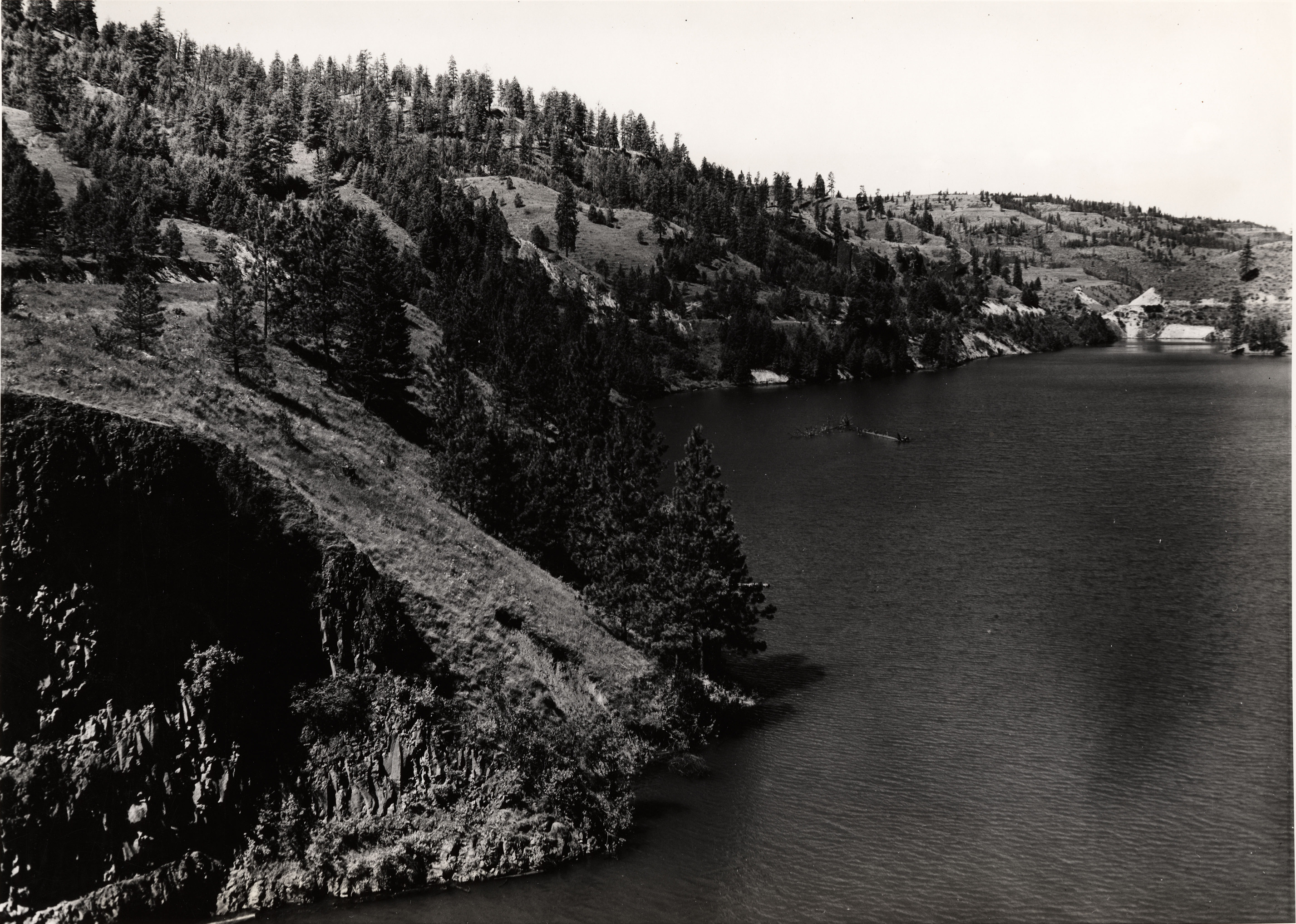 Black and white photograph of a body of water with a steep, forested shoreline