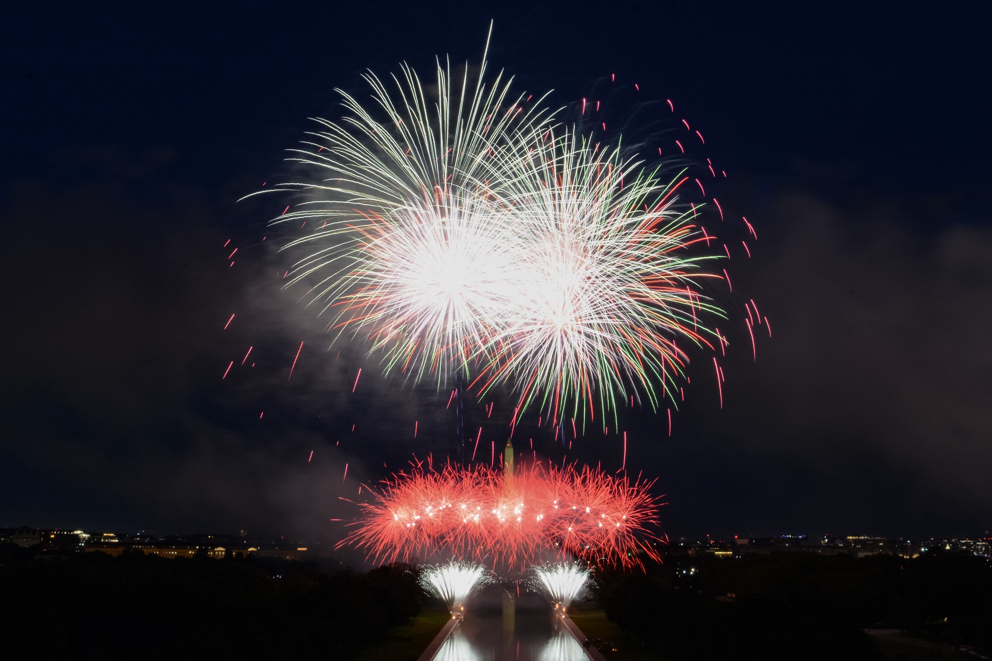 Colorful fireworks exploding and lighting up the dark sky above the Washington Monument and a reflecting pool.