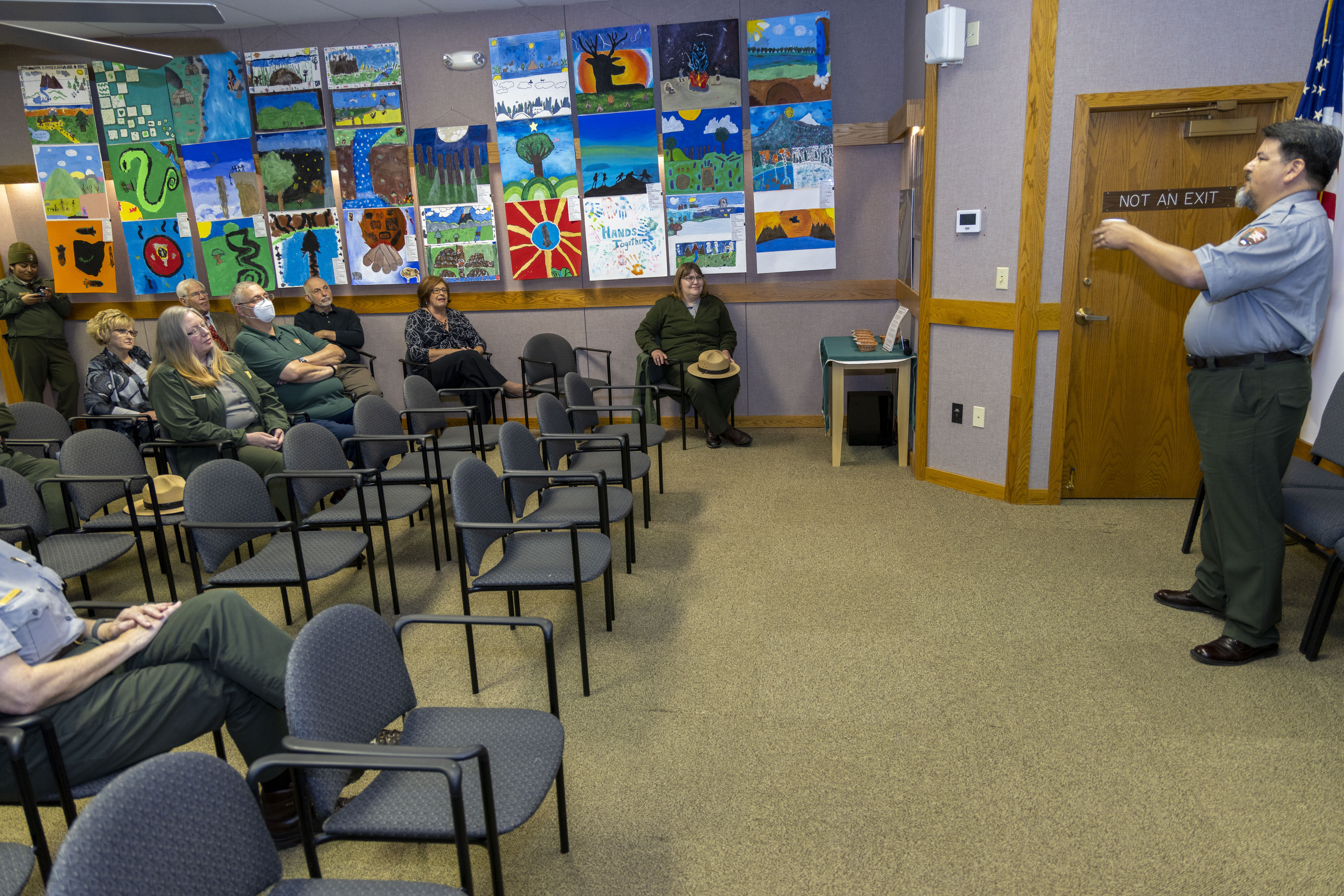 A man wearing a gray shirt and green pants speaks to a group of people sitting in chairs